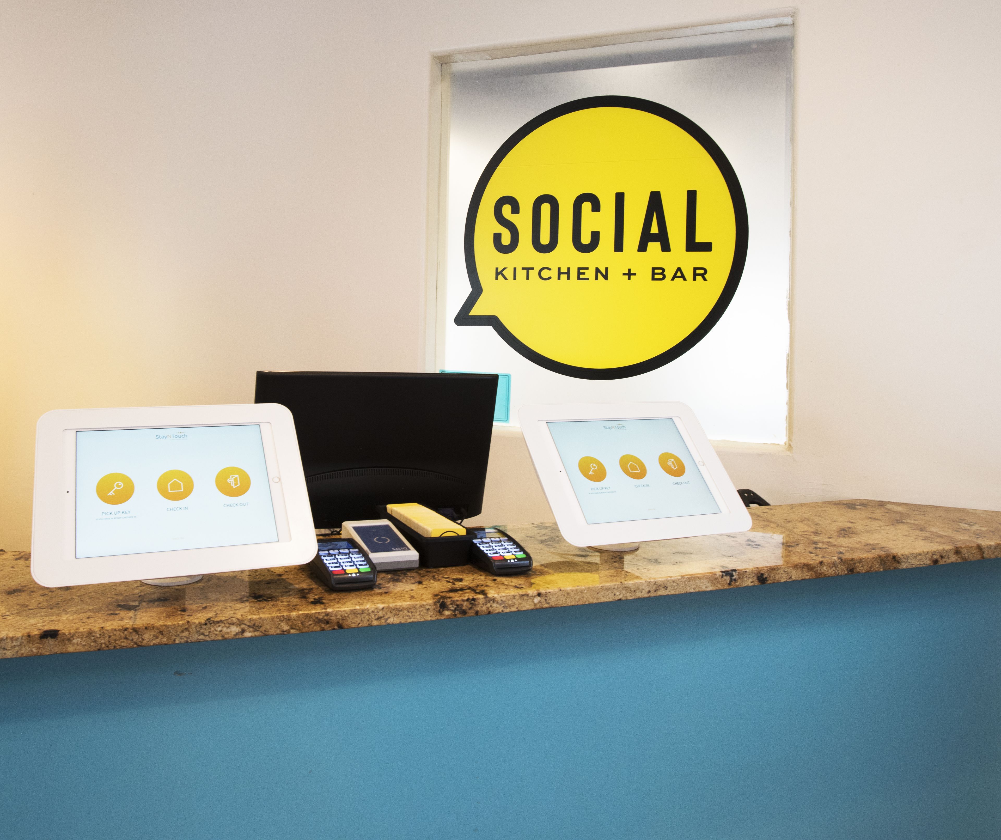 A counter with two tablets displaying a 'Stay-Touch' app interface, a cash register, and a 'Social Kitchen + Bar' sign.