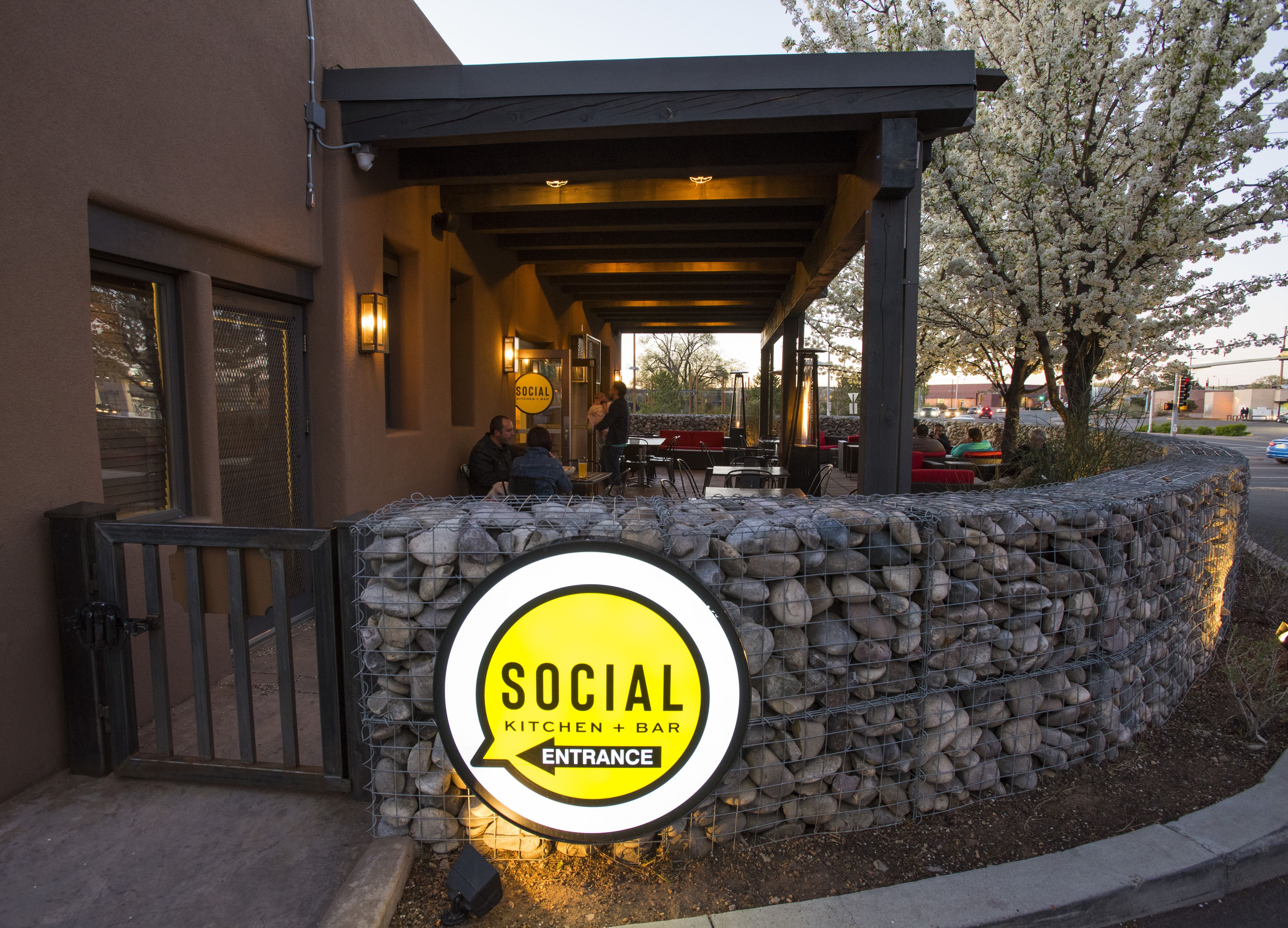 An outdoor seating area with tables and chairs under a covered patio, a stone wall with a sign reading "SOCIAL KITCHEN + BAR ENTRANCE", and a tree with white blossoms in the background.