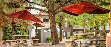Outdoor seating with red umbrellas and stone tables, surrounded by trees and gravel.