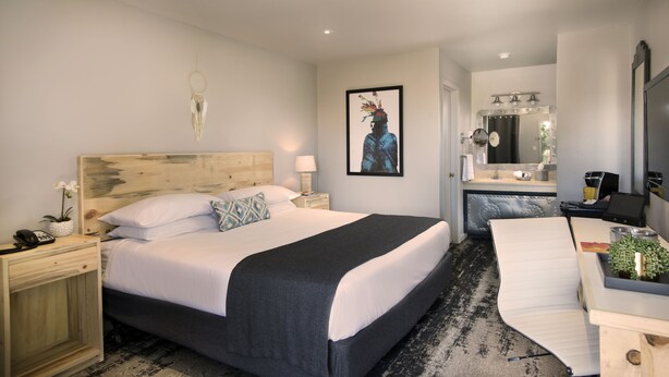 A modern hotel room with a large bed, bedside tables, a desk, and a bathroom with a mirror and sink.