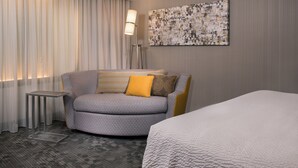 Habitación de hotel con un sofá gris, una mesita auxiliar pequeña, una lámpara, un cuadro abstracto grande y una cama con colcha blanca.