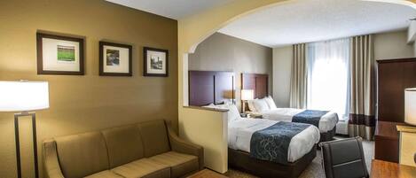 A hotel room with a sofa, two beds, a desk, and framed pictures on the wall.