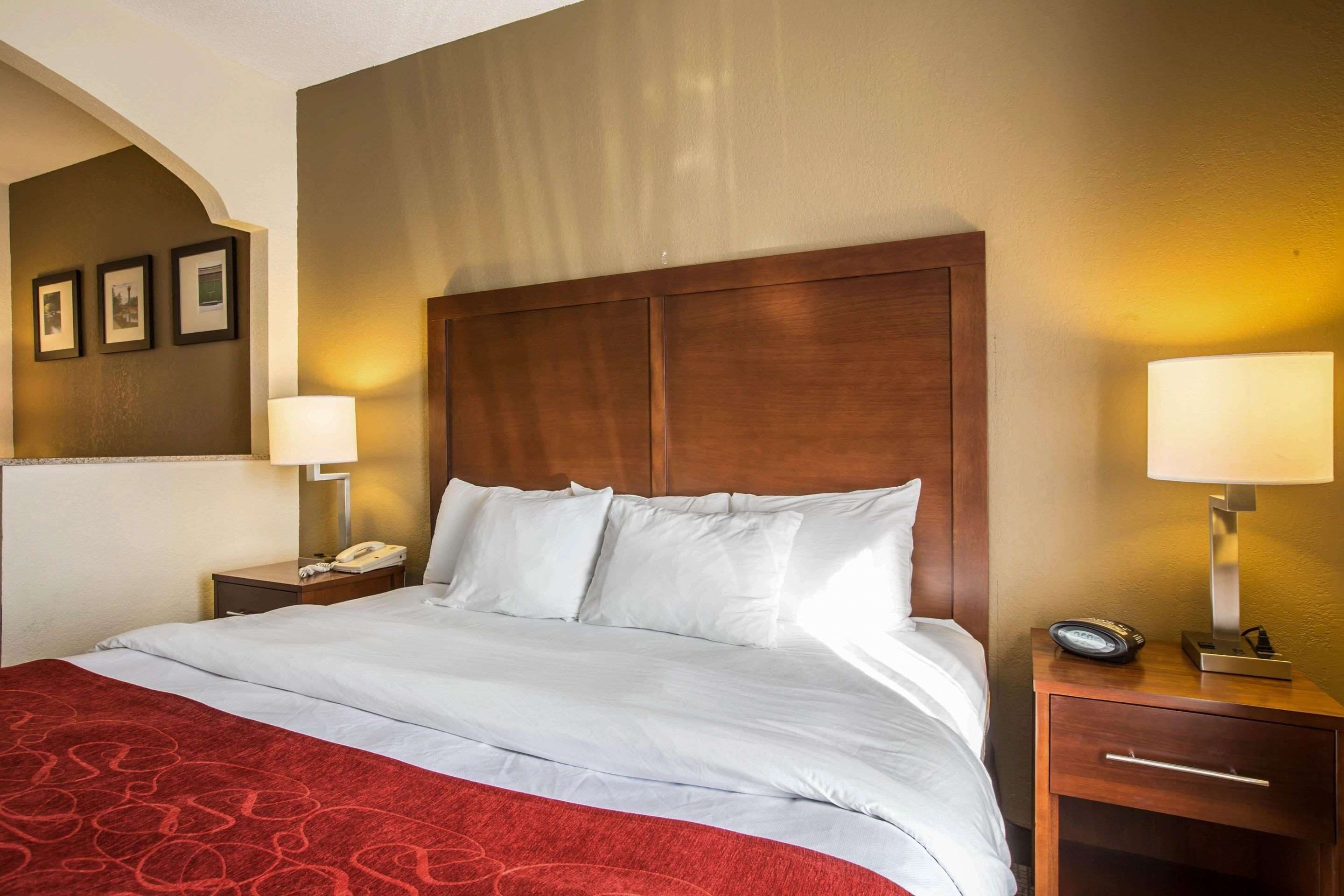 A hotel room with a large bed, two bedside tables, a wooden headboard, and a red patterned bedspread.