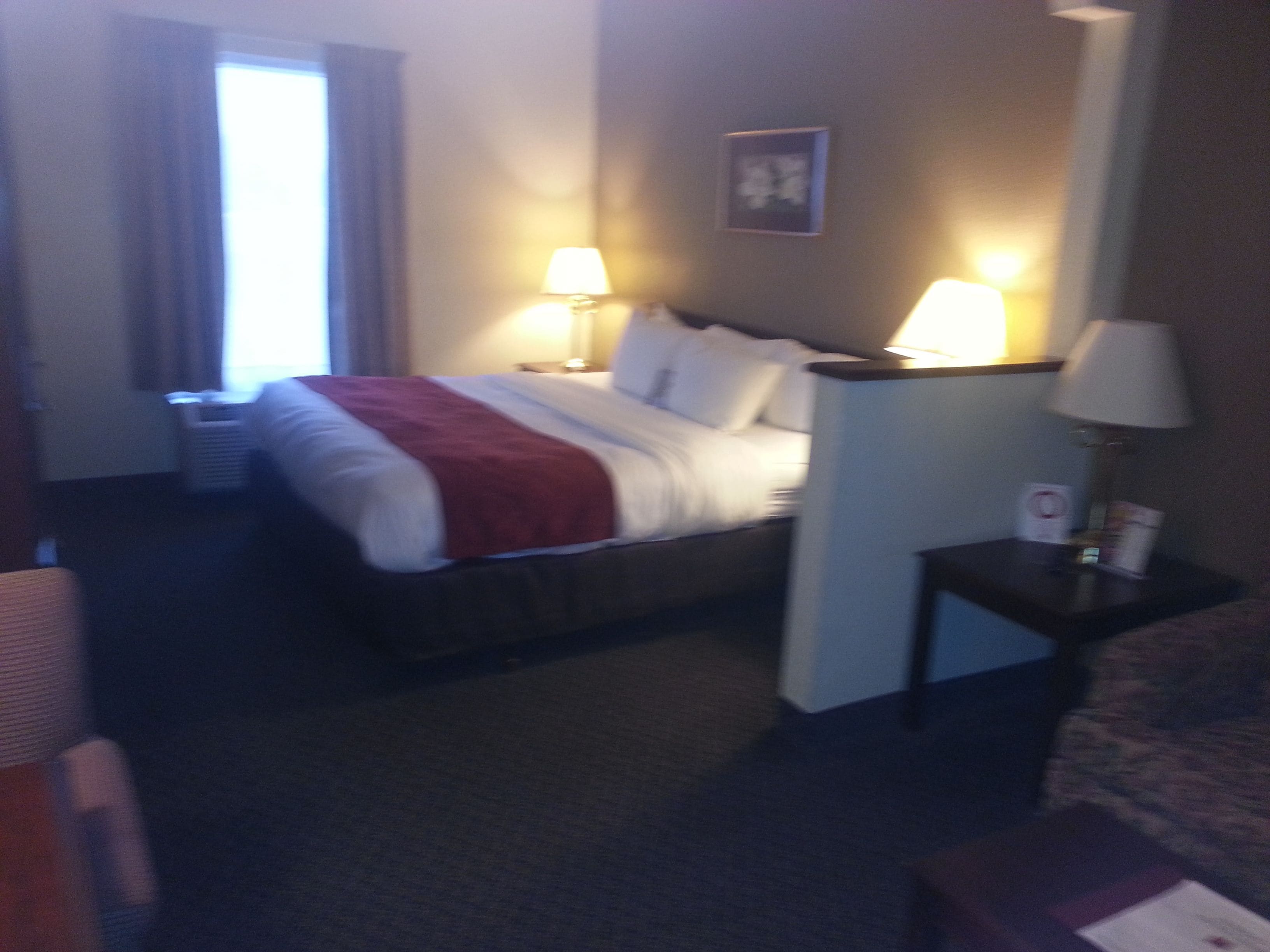 A hotel room with a bed, two bedside lamps, a nightstand, a window with curtains, and a wall-mounted picture.