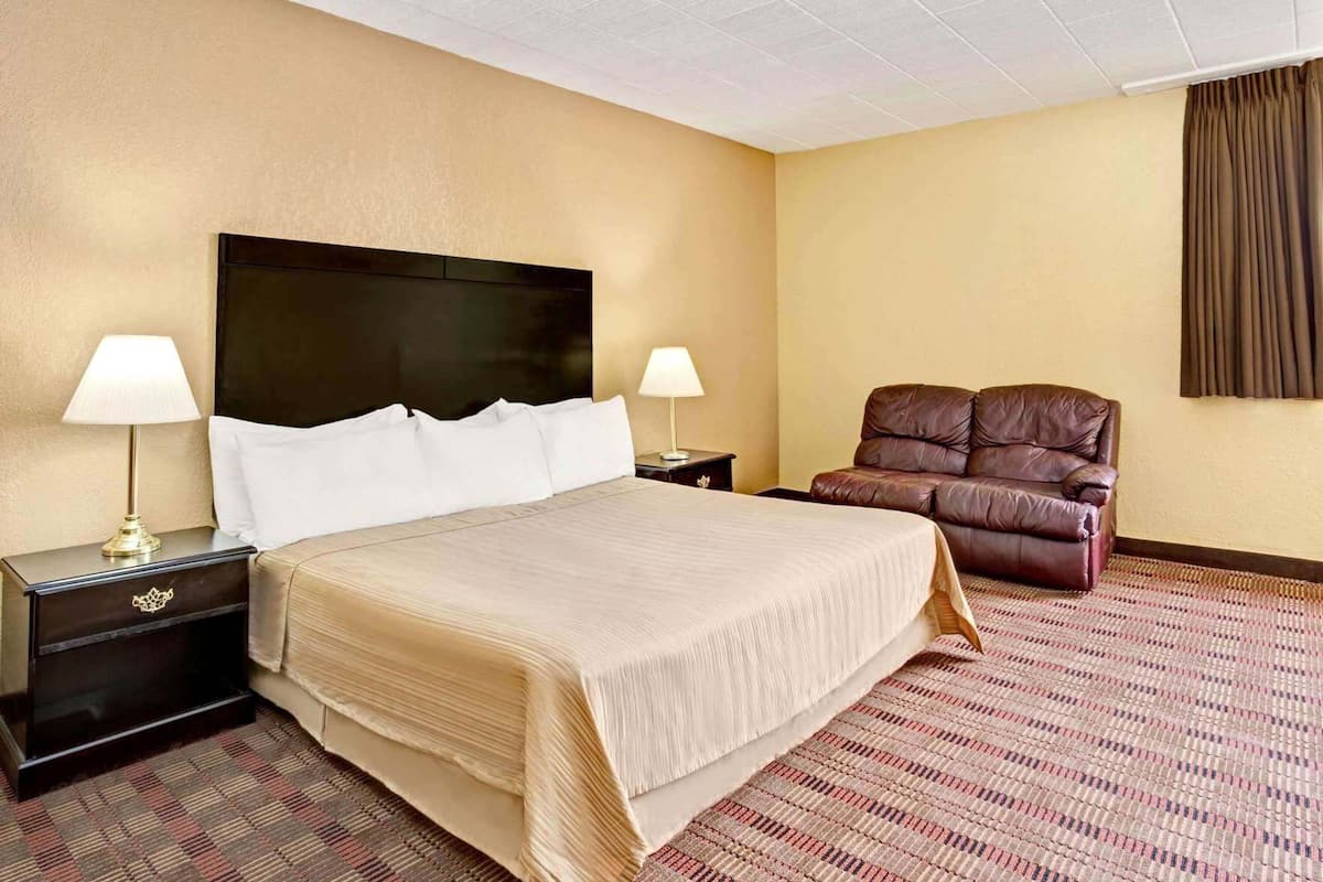 A hotel room with a large bed, two bedside tables with lamps, a leather armchair, and a plain wall.