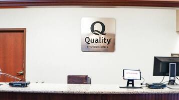Un comptoir d’accueil avec une enseigne « Qualité par Choice Hotels », un panneau d’accueil, un écran d’ordinateur et une horloge murale.