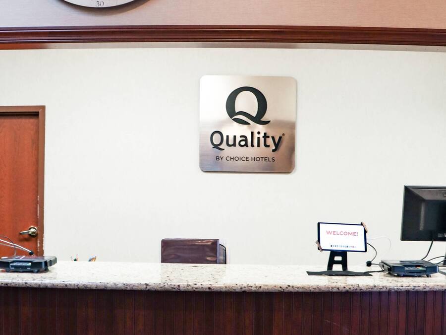 Un comptoir d’accueil avec une enseigne « Qualité par Choice Hotels », un panneau d’accueil, un écran d’ordinateur et une horloge murale.