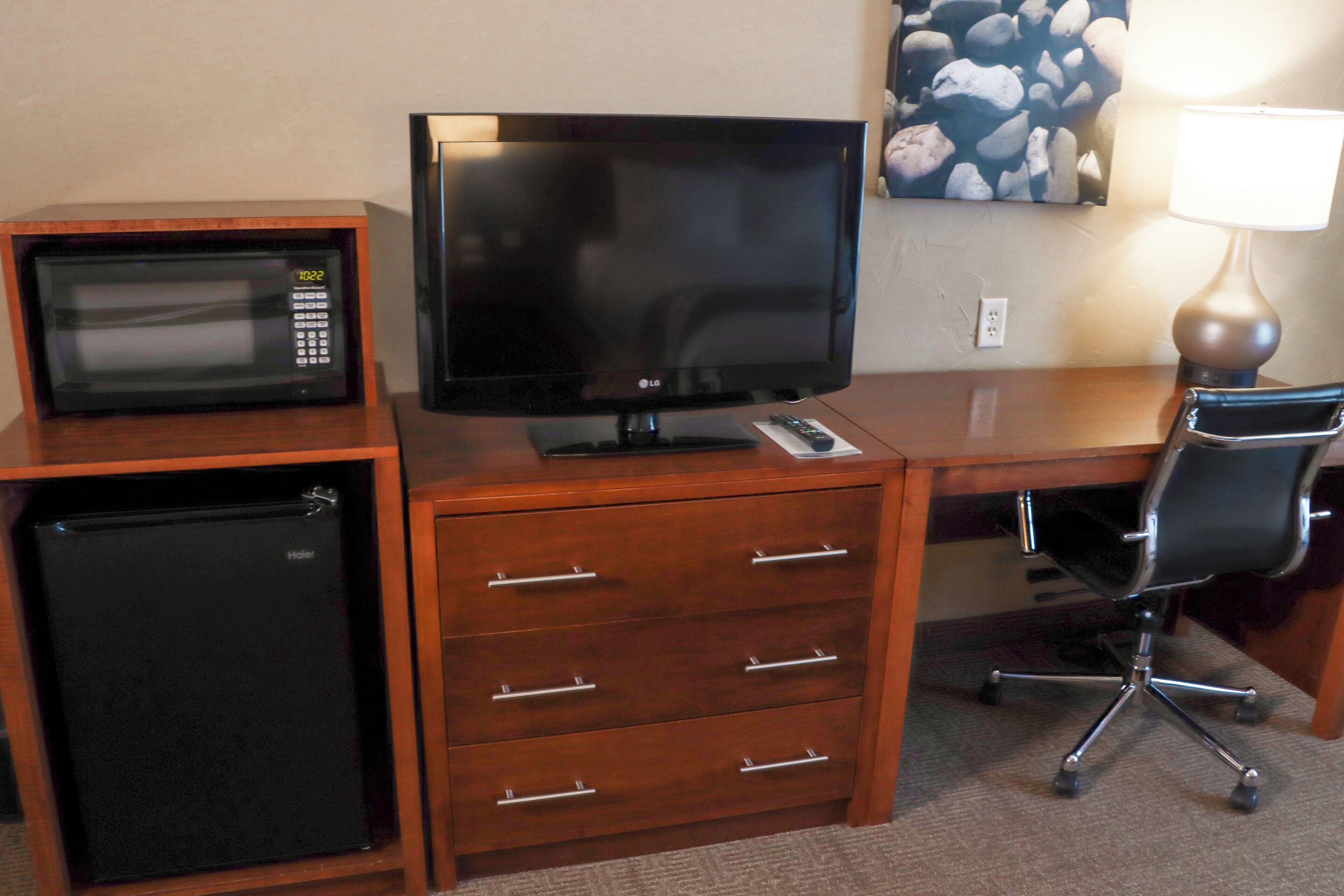 A compact kitchenette with a microwave, refrigerator, and a flat-screen TV on a wooden desk.