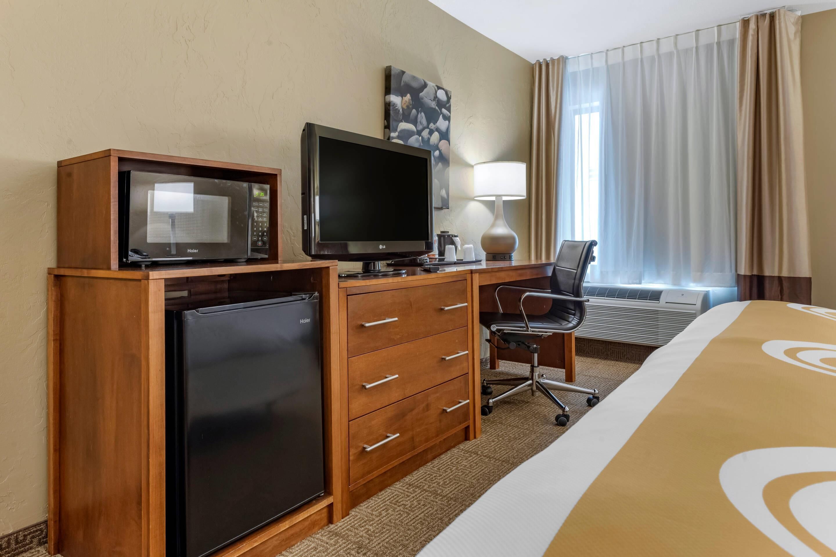 A hotel room with a bed, a desk with a chair, a microwave, a refrigerator, a television, a lamp, and a window with curtains.