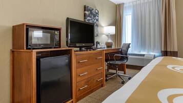 A hotel room with a bed, a desk with a chair, a microwave, a refrigerator, a television, a lamp, and a window with curtains.