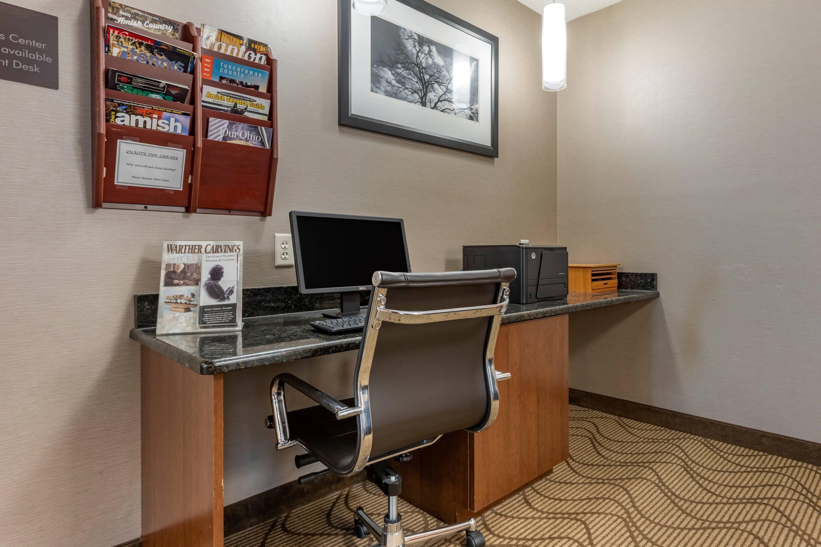 Bancada de recepção de hotel com um computador, uma cadeira, um quadro emoldurado e um mural com diversos avisos.