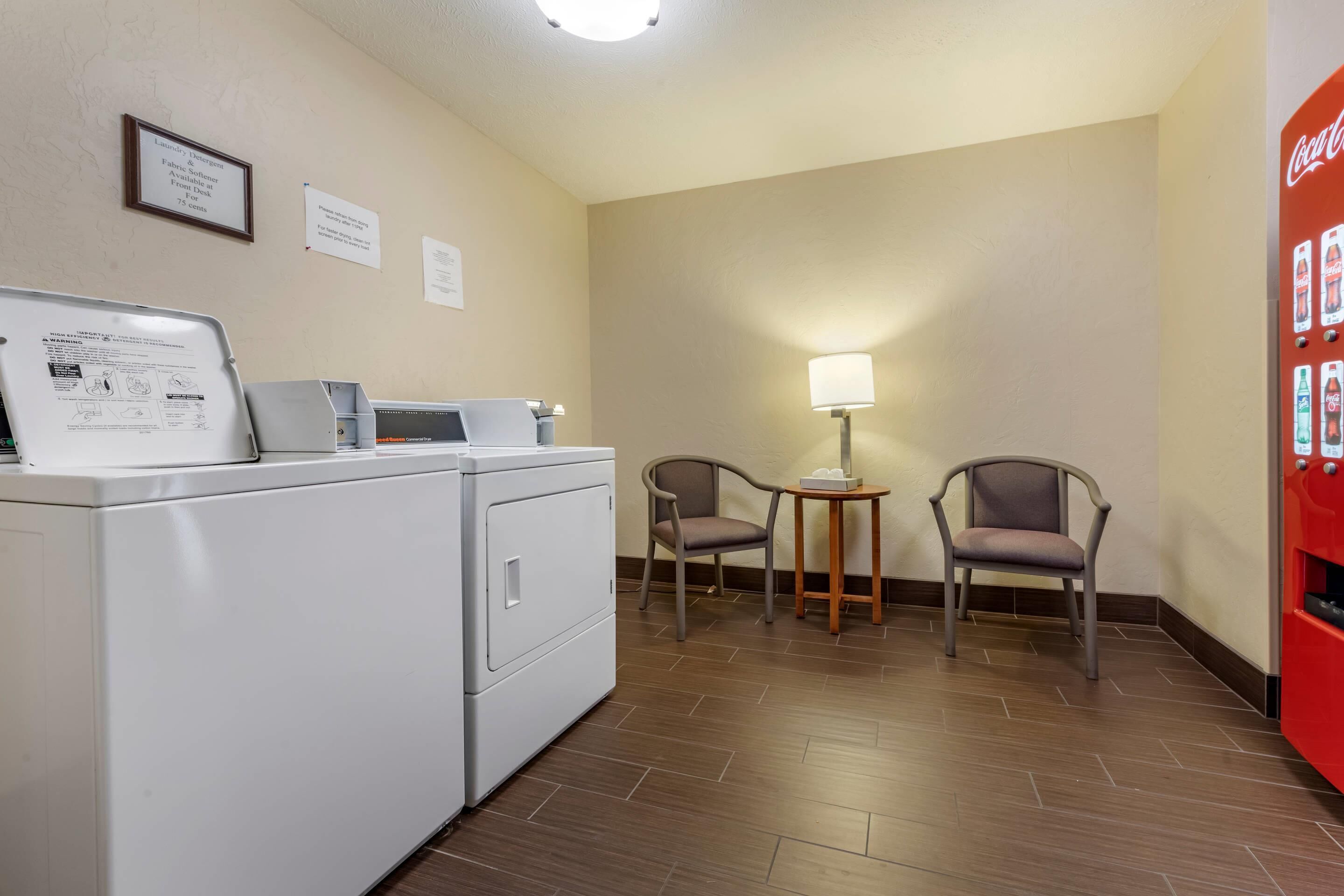 Habitación de hotel con lavadora, dos sillas, una mesita con lámpara y refrigerador Coca-Cola.