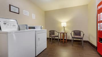 Habitación de hotel con lavadora, dos sillas, una mesita con lámpara y refrigerador Coca-Cola.