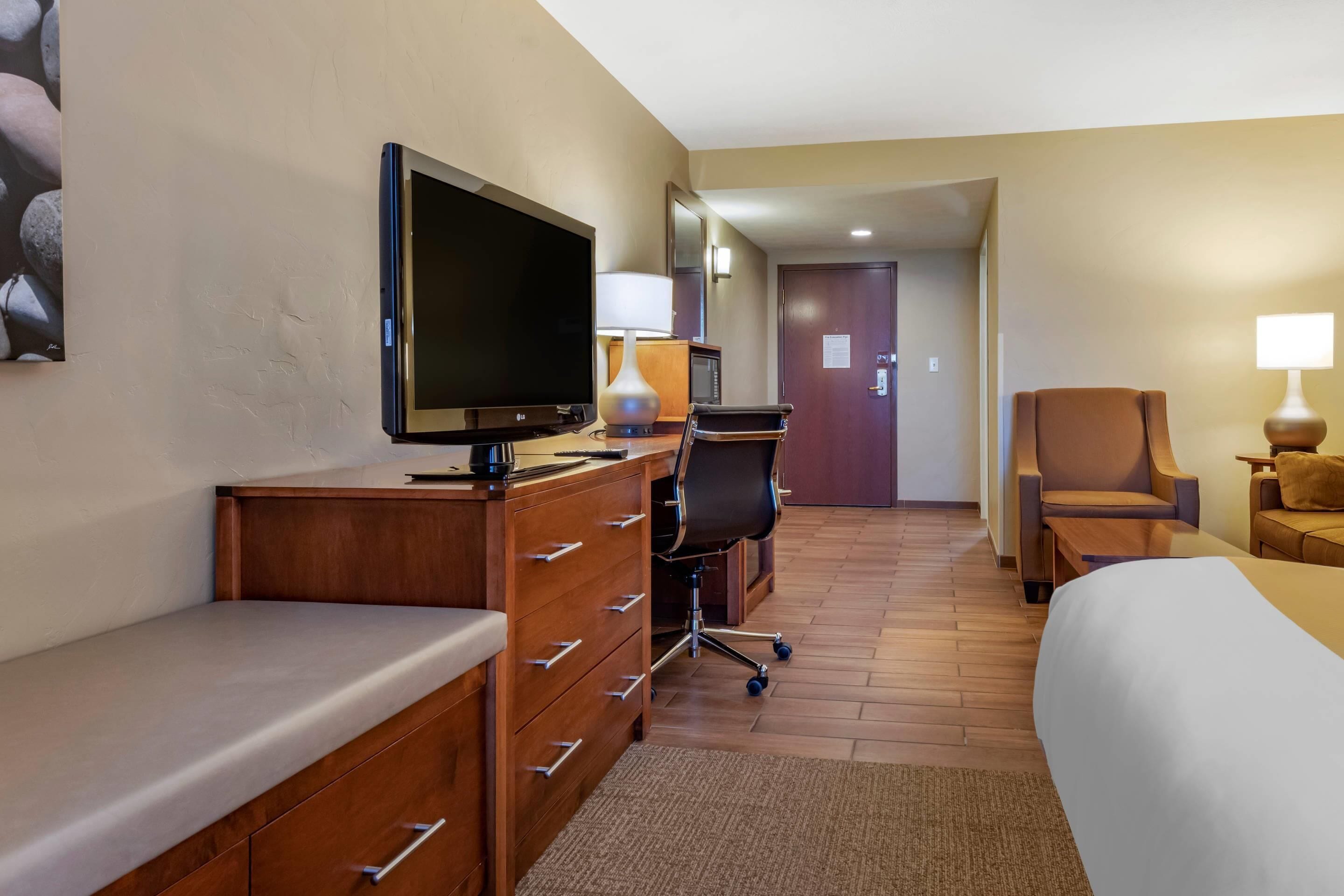 Una habitación de hotel con cama, escritorio con televisor, silla, lámpara de escritorio y puerta.