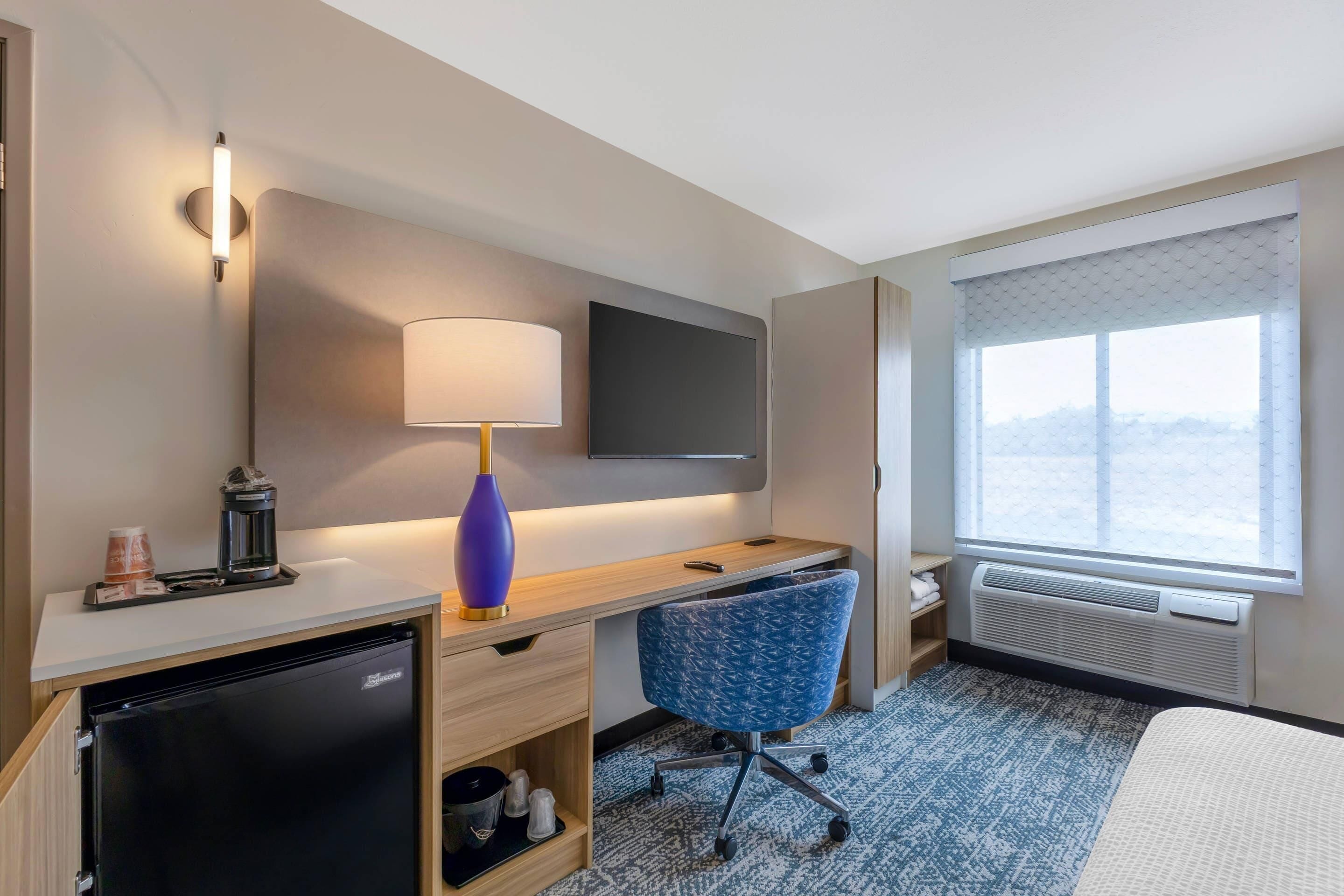 Une chambre d’hôtel équipée d’un bureau, d’une chaise, d’une télévision, d’un mini-réfrigérateur et d’une grande fenêtre.