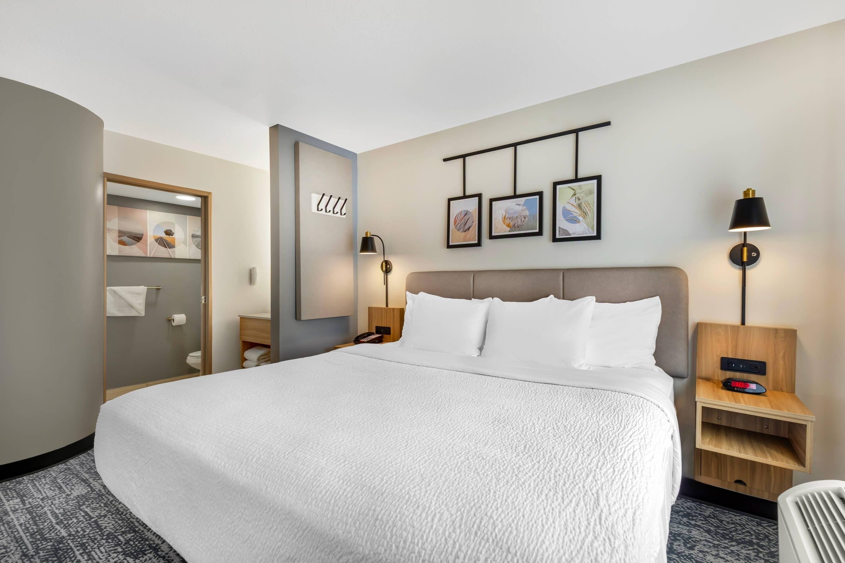 A hotel room with a large bed, a bedside table with a lamp, a wall-mounted clock, and framed artwork.