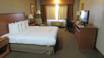 A hotel room with a large bed, a desk with a television, a chair, an air conditioning unit, and a lamp.