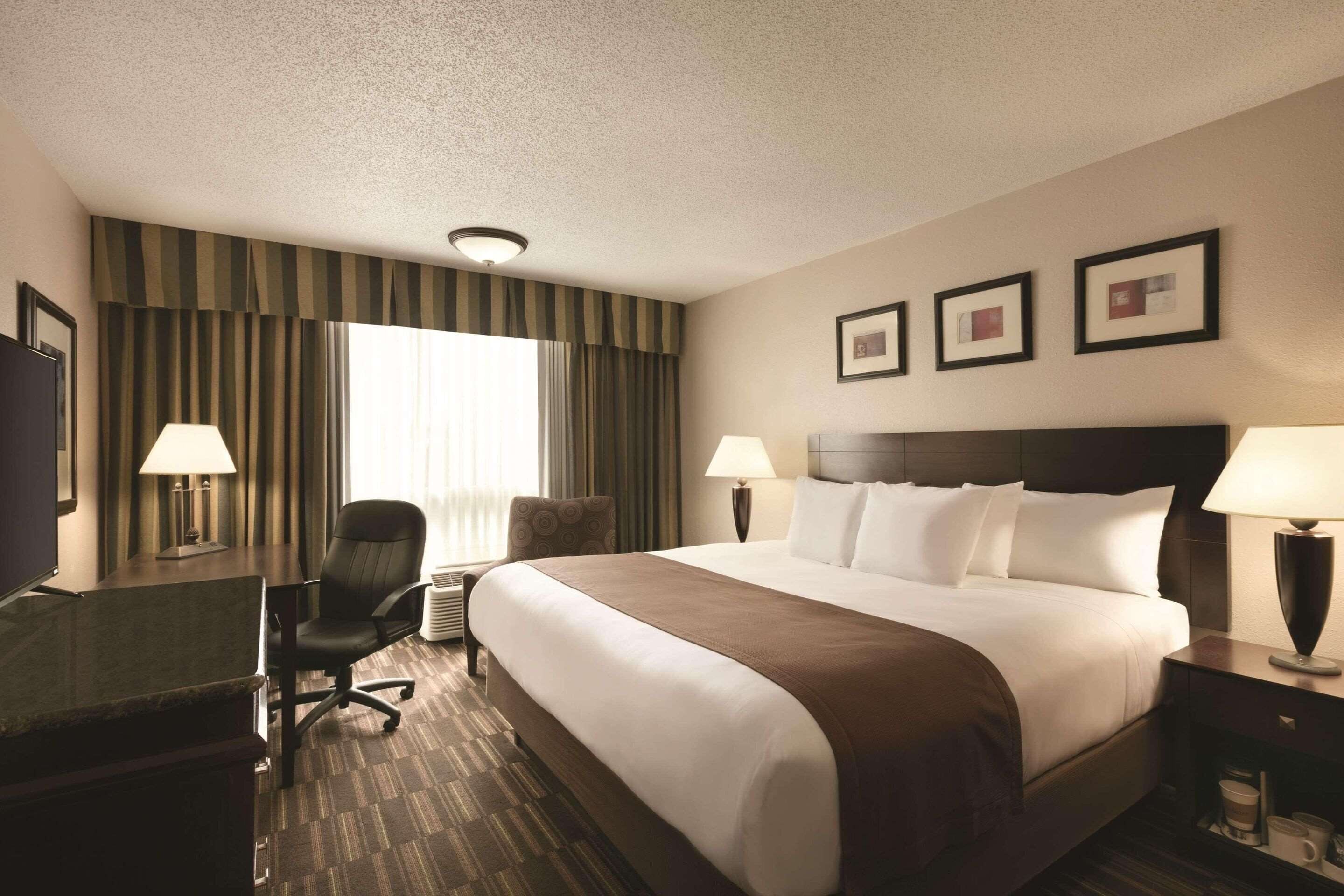 A hotel room with a large bed, two bedside lamps, a desk with a lamp, a chair, and a large window with curtains.