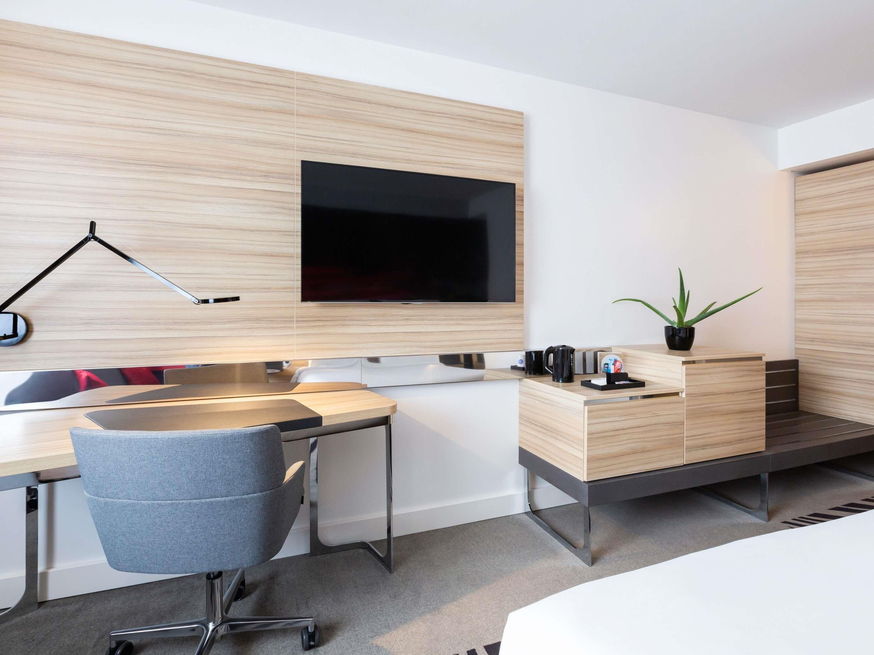 Habitación de hotel moderna con escritorio, silla, televisor de pantalla plana, mesita de noche y una planta en maceta.