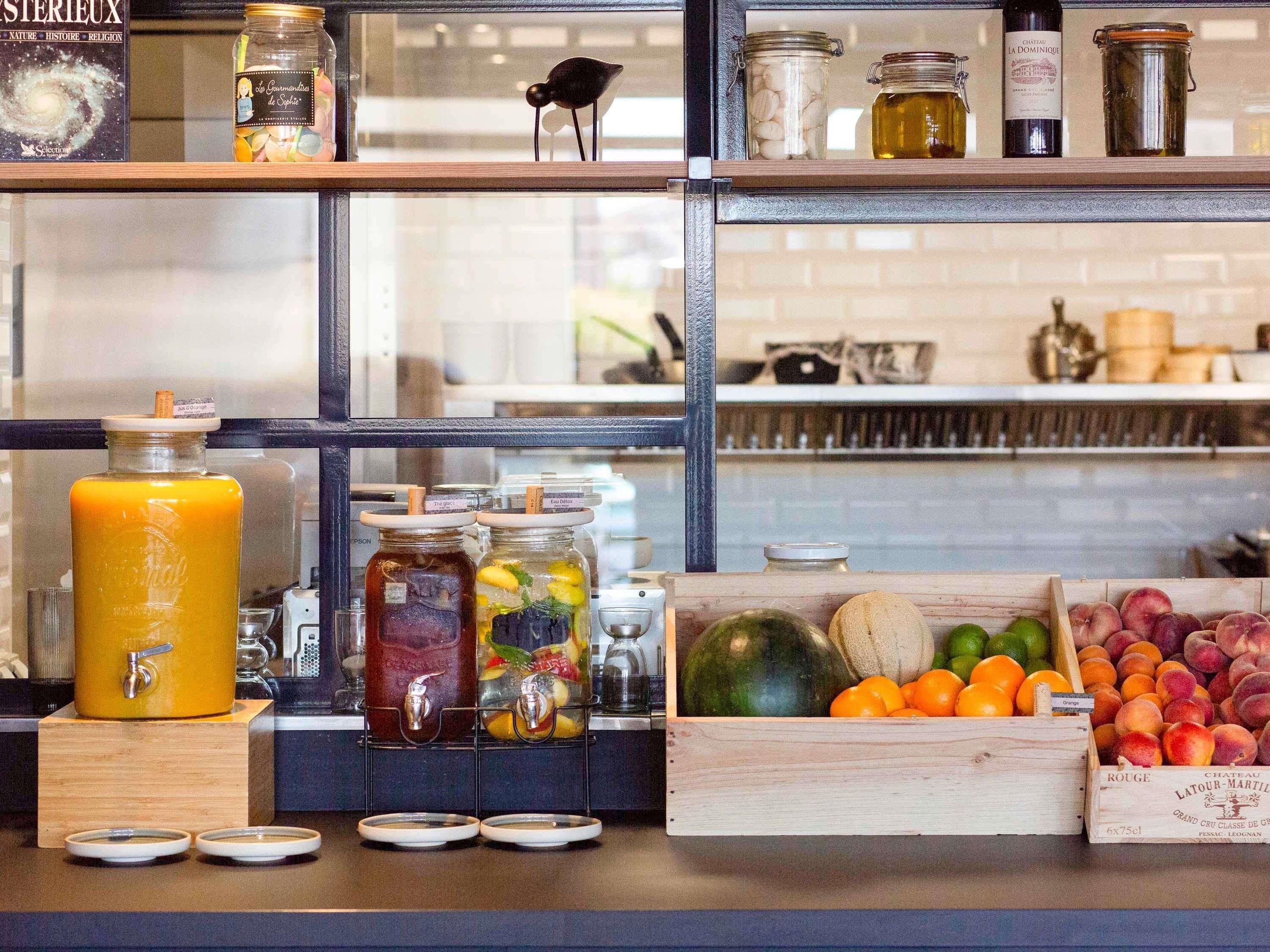 Een drankstation met een grote gele container, meerdere glazen potten met rietjes, en een houten krat vol fruit.