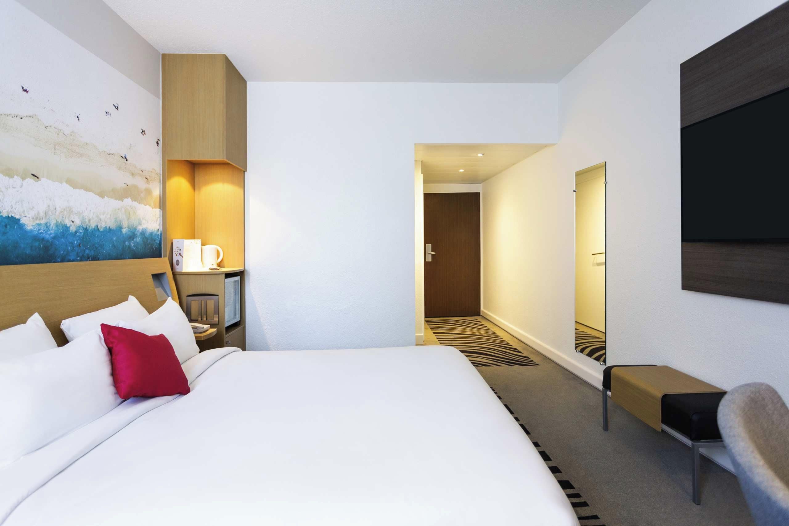 A hotel room with a large bed, a desk with a chair, a painting on the wall, and a view into the hallway.
