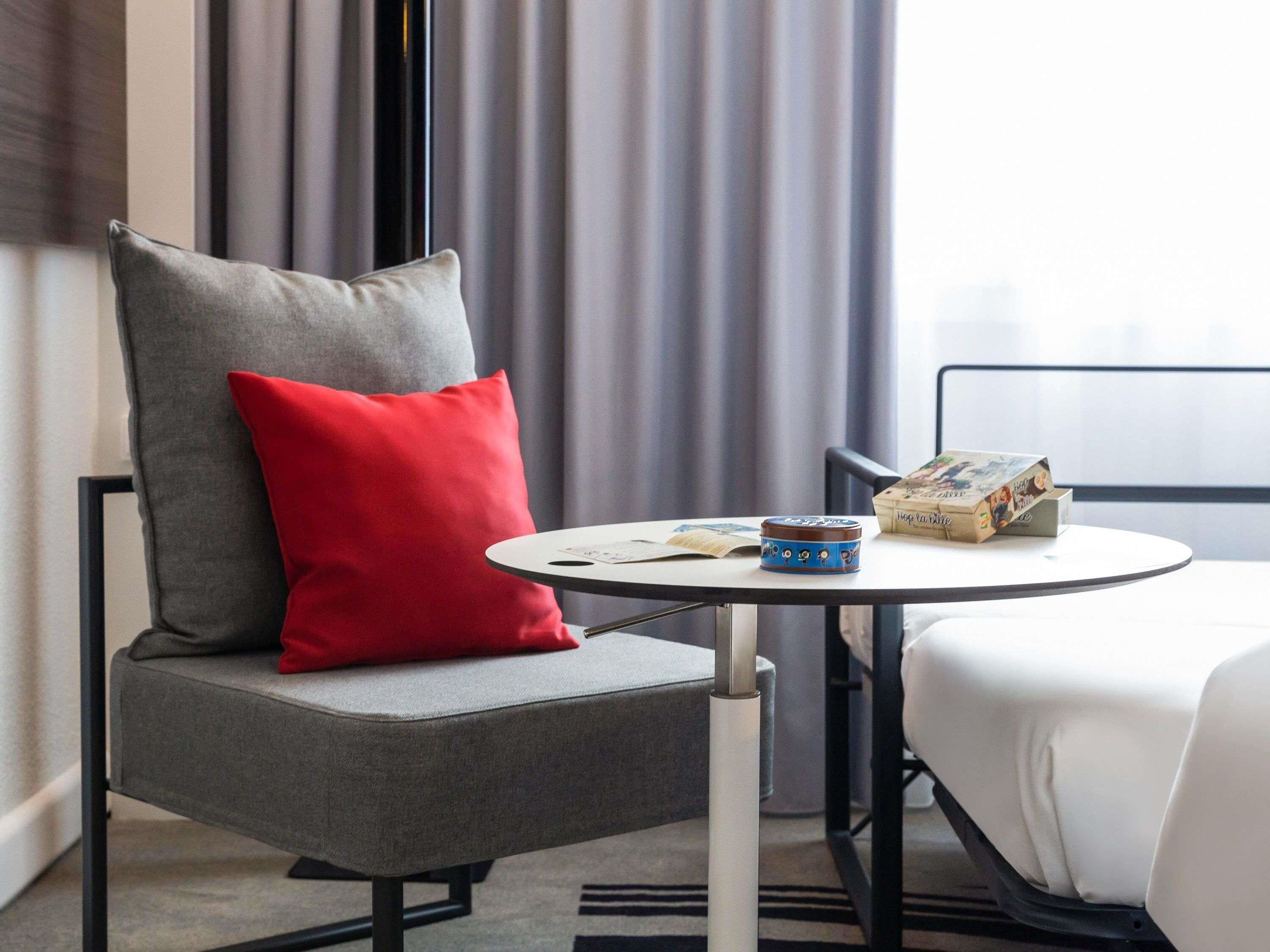 Habitación de hotel con una silla tapizada de gris, un cojín rojo, una mesita redonda con una taza azul y un libro, y una cama blanca con estructura negra.