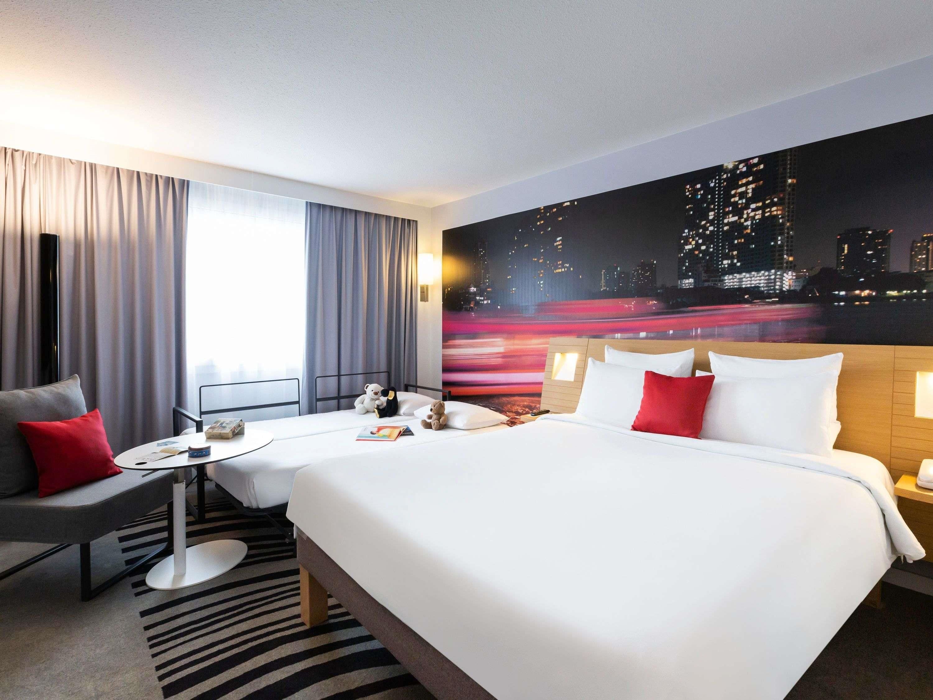 A hotel room with a bed, a desk, a chair, a window with curtains, and a cityscape view on the wall.
