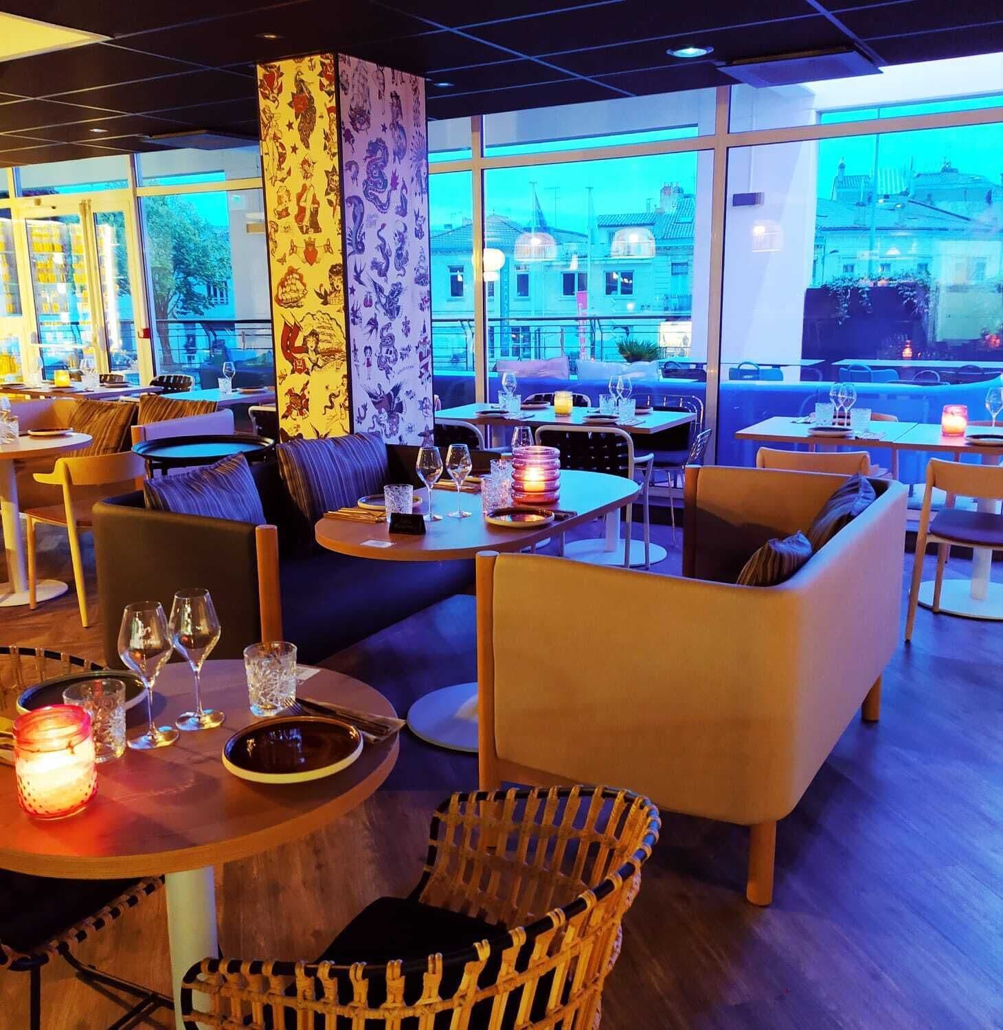 Um restaurante moderno com mesas redondas, assentos acolchoados e amplas janelas com vista para a cidade.