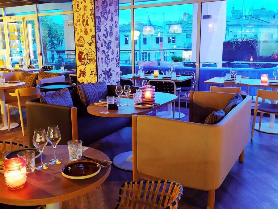 Um restaurante moderno com mesas redondas, assentos acolchoados e amplas janelas com vista para a cidade.