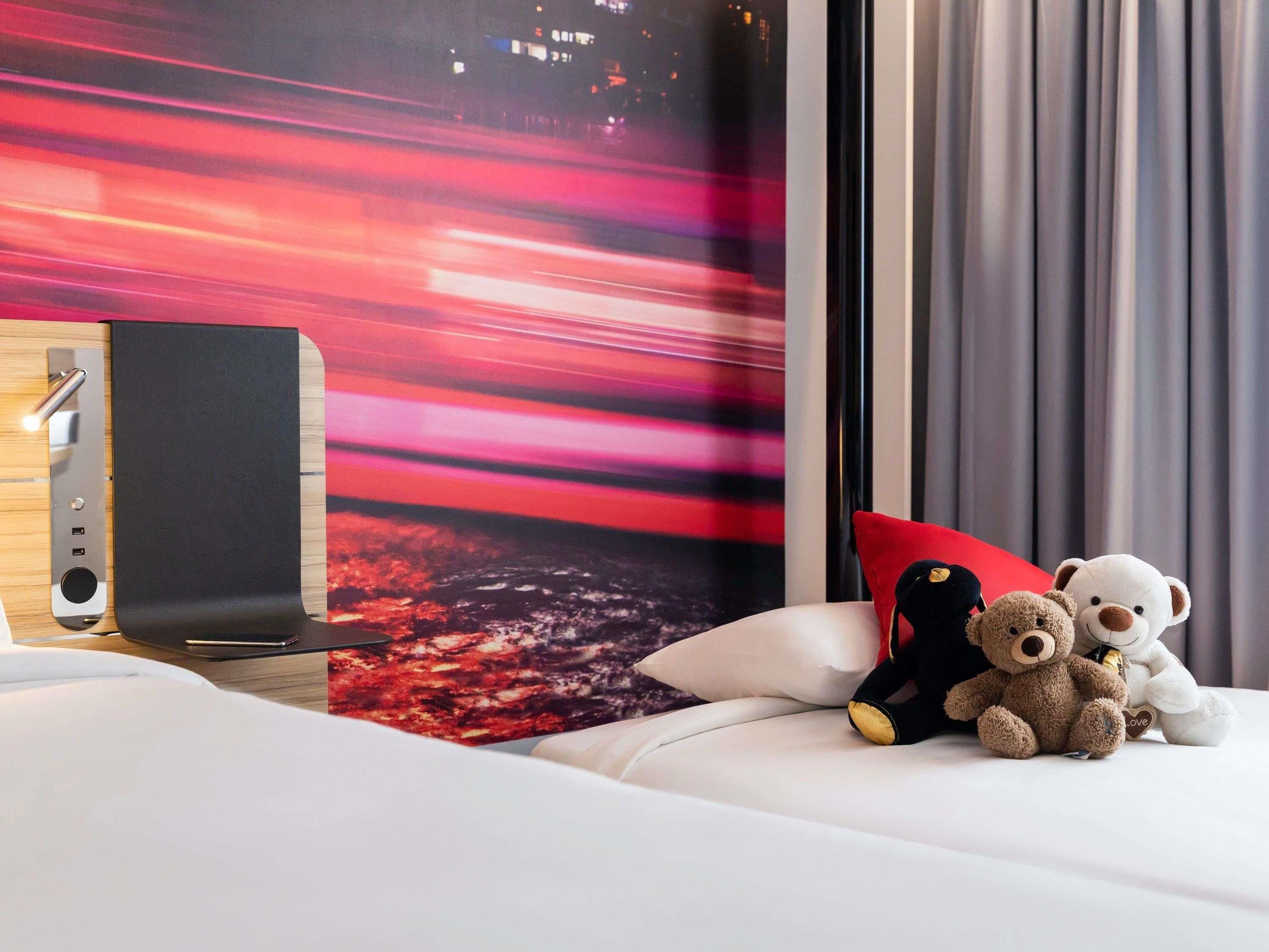 Quarto de hotel com uma cama, um alto-falante fixado na parede e dois ursinhos de pelúcia na cama.