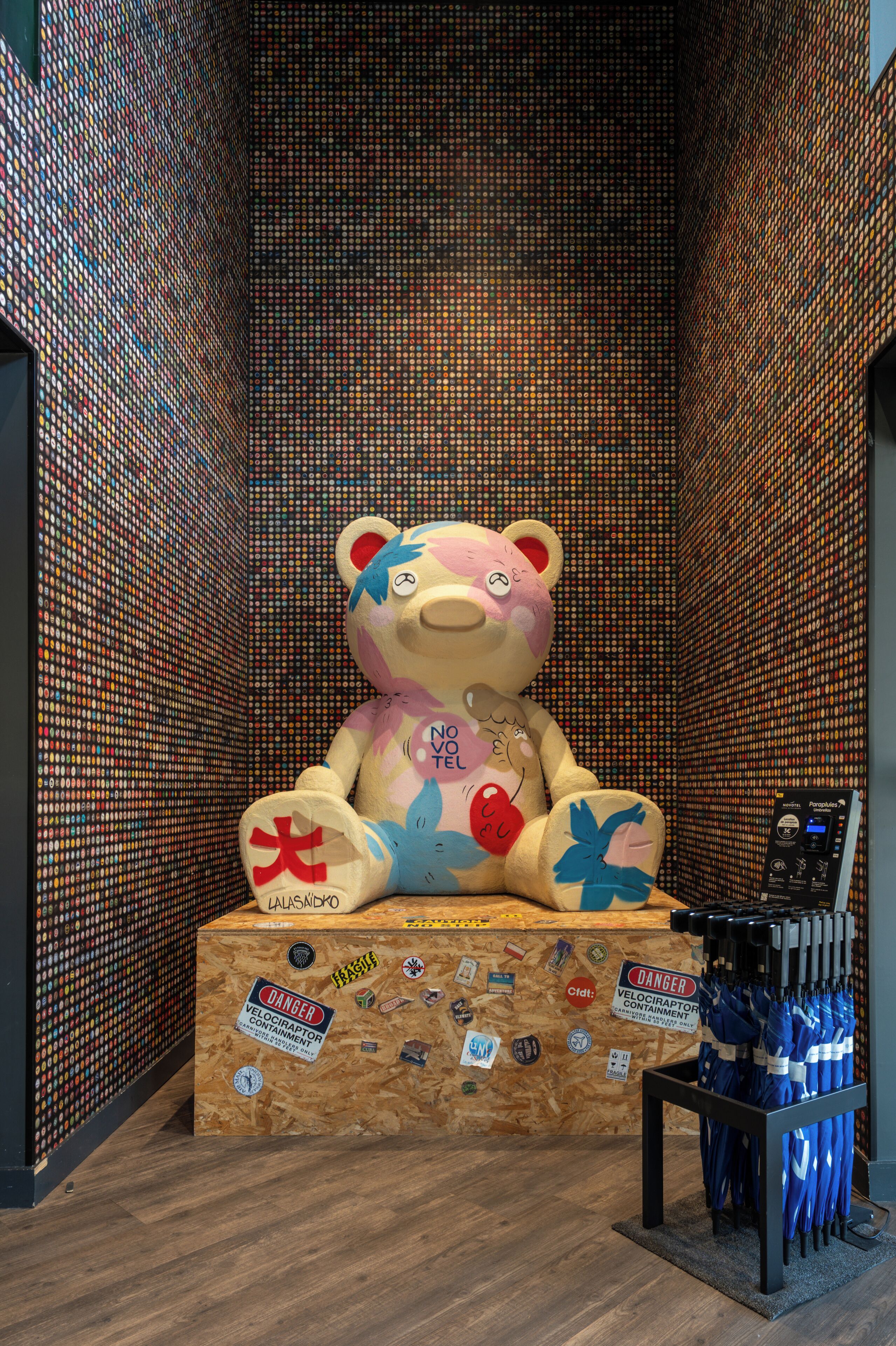 Eine Bärenskulptur mit Aufklebern auf einer Holzplattform, umgeben von einer Wand aus bunten Kronkorken.