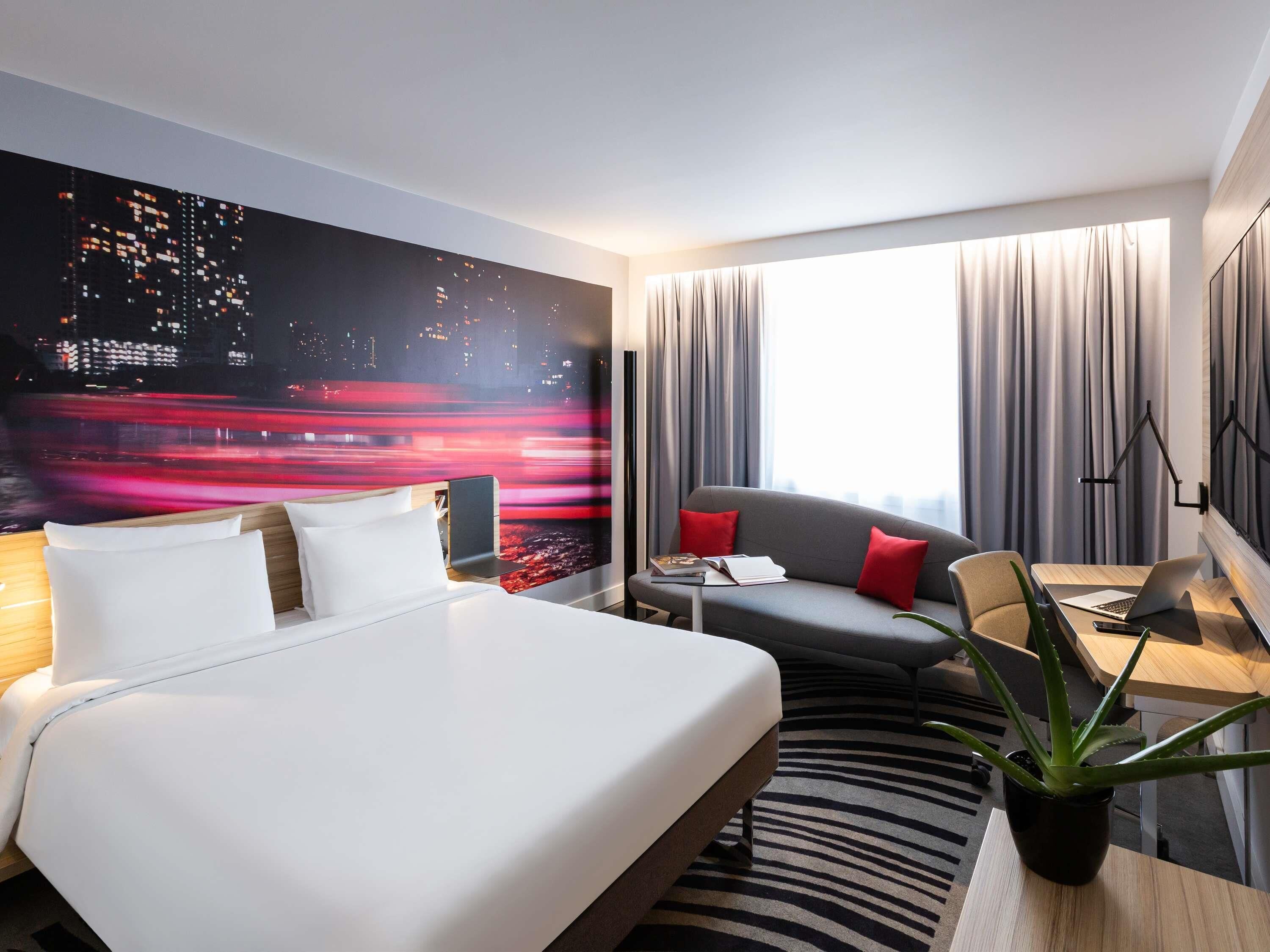 Hotelkamer met een bed, een slaapbank, een bureau met een laptop, een plant en een groot raam met gordijnen.