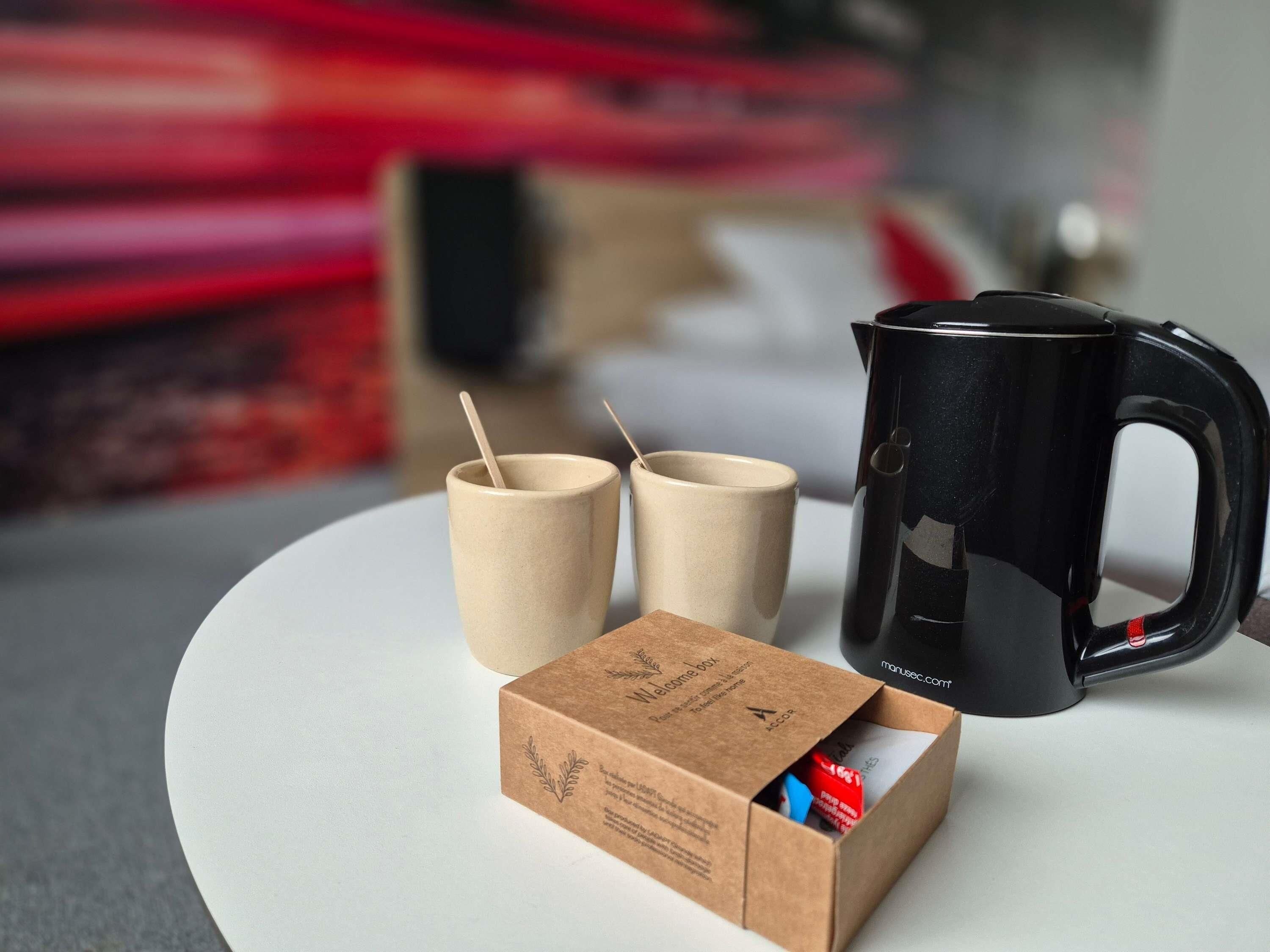 A black coffee maker, two ceramic cups, and a cardboard box with a logo on a white surface.