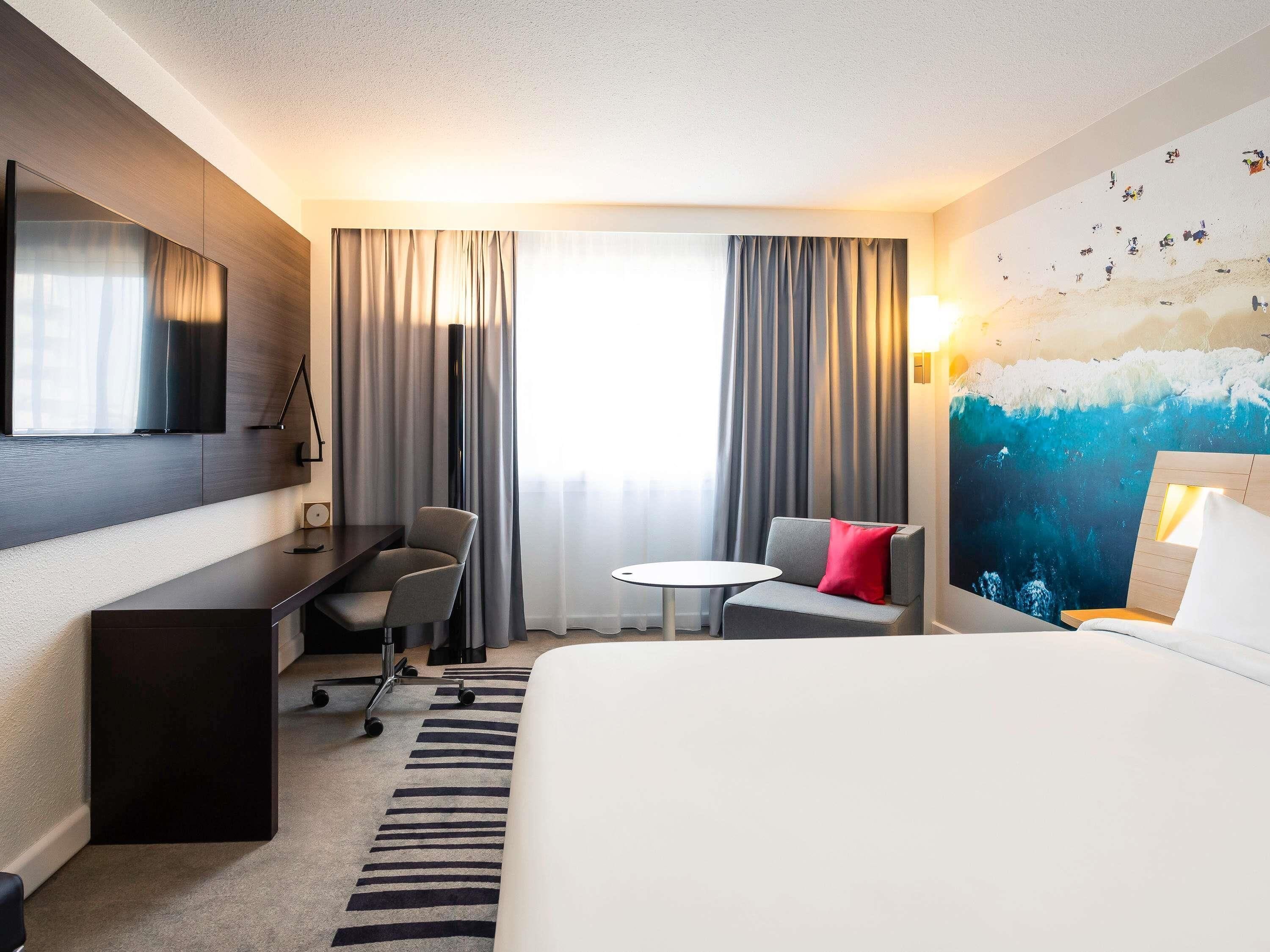 Habitación de hotel con cama, escritorio, silla, mesita y un ventanal grande con cortinas.