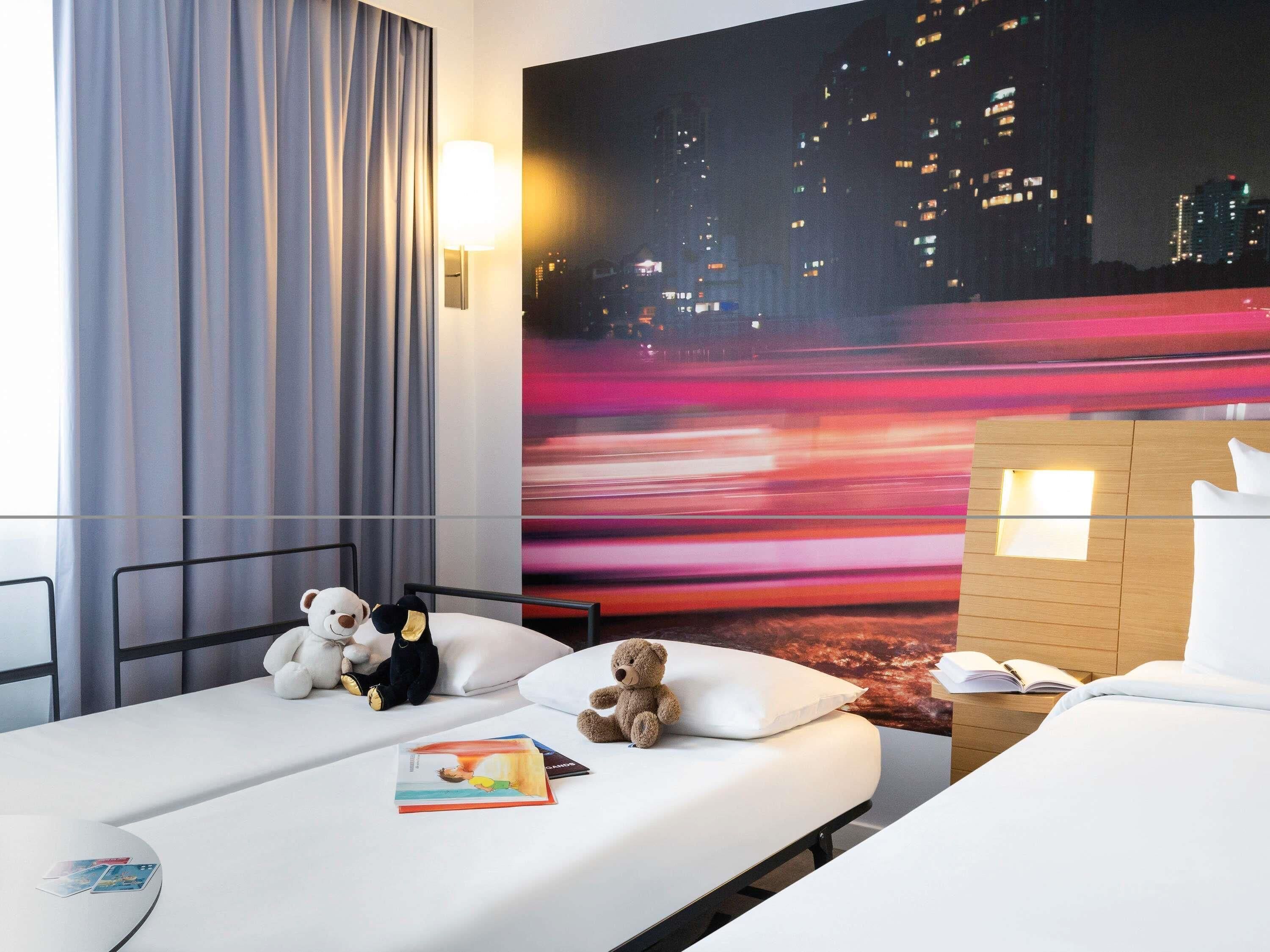 A hotel room with a bed, a bedside table, a lamp, a teddy bear, and a cityscape mural on the wall.