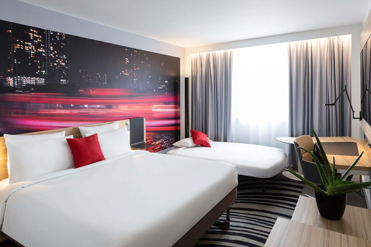 A hotel room with a bed, a desk, a chair, a plant, and a cityscape mural on the wall.