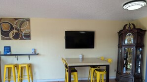 A dining area with a table and yellow chairs, a flat-screen TV, a clock, and a shelf with a sign.