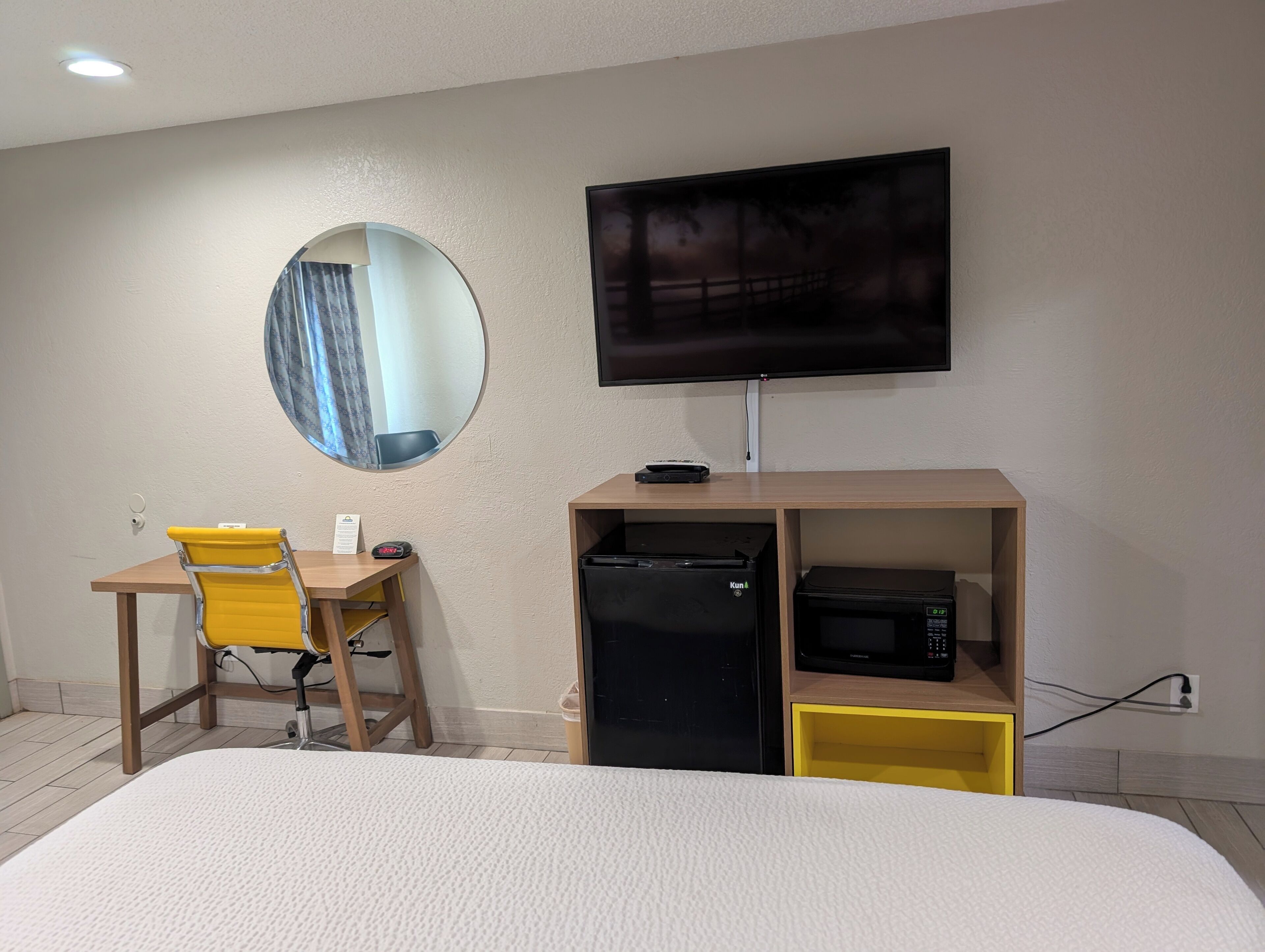 A hotel room with a bed, a desk with a chair, a television, a minibar, and a mirror.