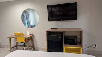 Una habitación de hotel con cama, escritorio con silla, televisor, minibar y espejo.