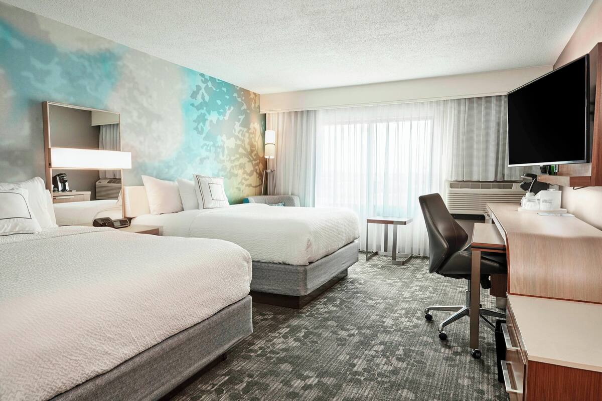 Une chambre d’hôtel avec deux lits, un bureau, une chaise, une télévision et une grande fenêtre avec des rideaux.