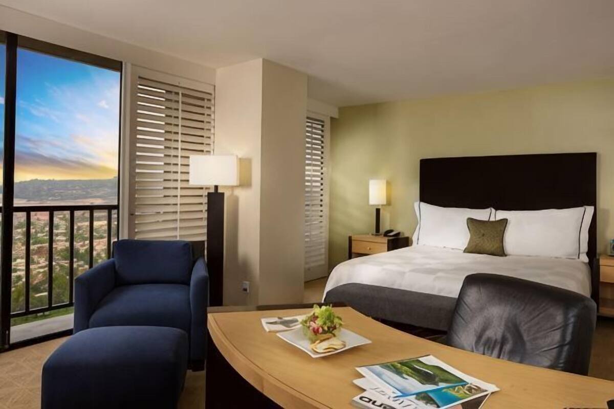 Habitación de hotel con cama, escritorio y silla, mesa con una revista, lámpara y vista por la ventana.