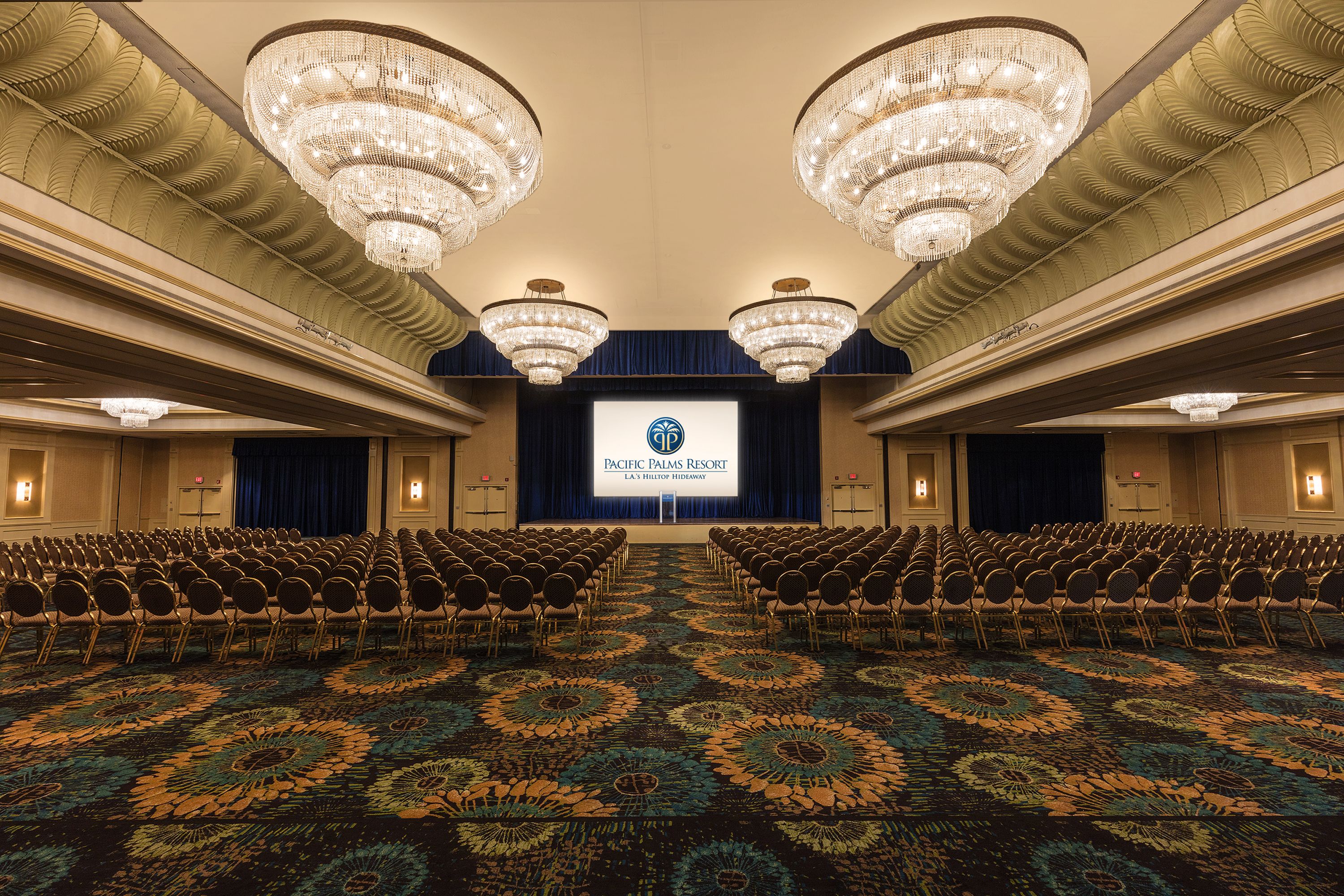 A large conference room with rows of chairs facing a stage, chandeliers, and a floral carpet.