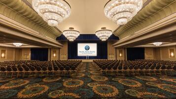 A large conference room with rows of chairs facing a stage, chandeliers, and a floral carpet.