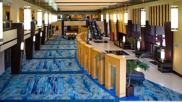A hotel lobby with a blue patterned carpet, wooden reception desk, escalators, and a large potted plant.