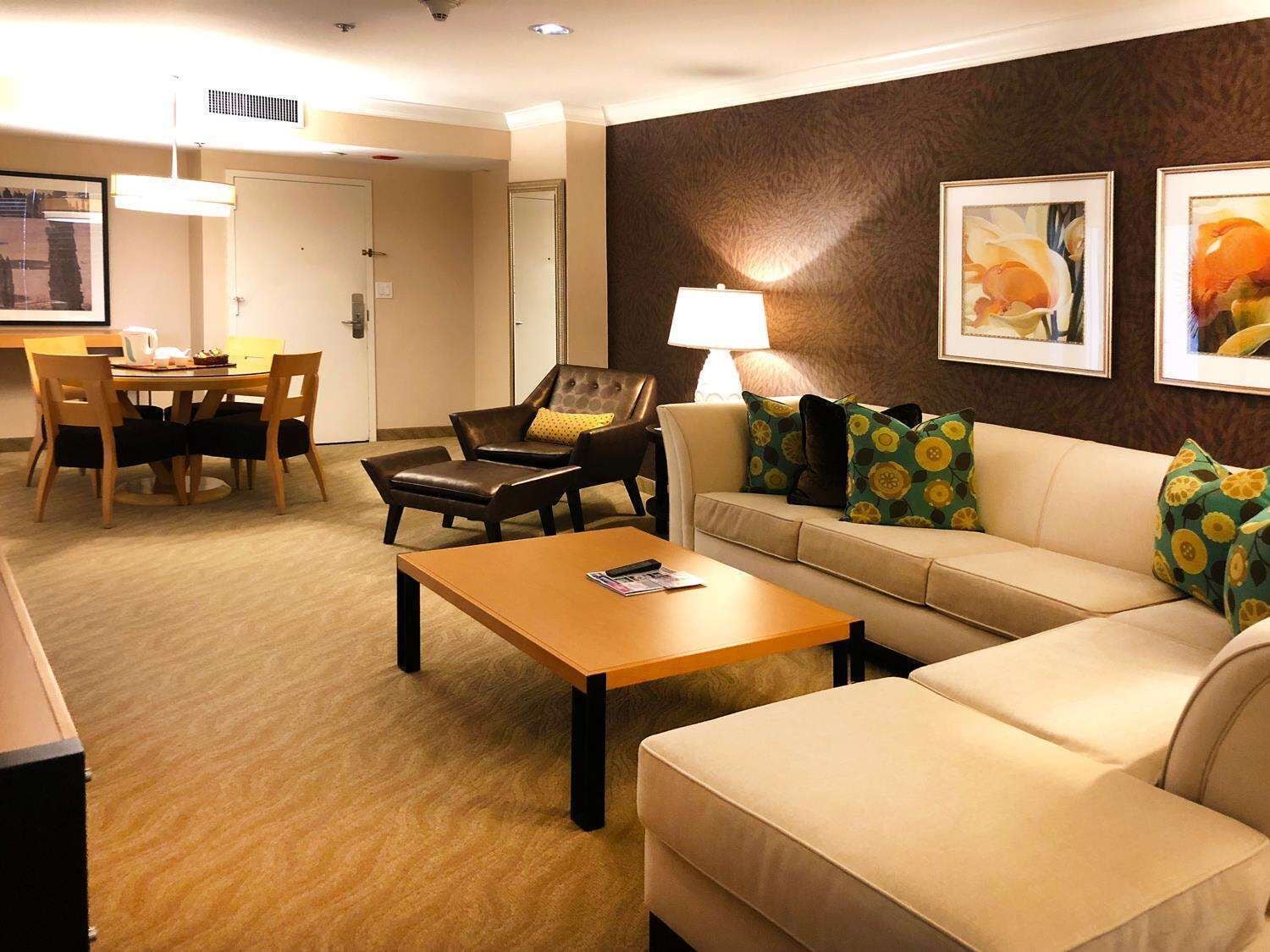 Habitación de hotel con sofá, sillón, mesa de centro, mesa de comedor, sillas y lámpara.