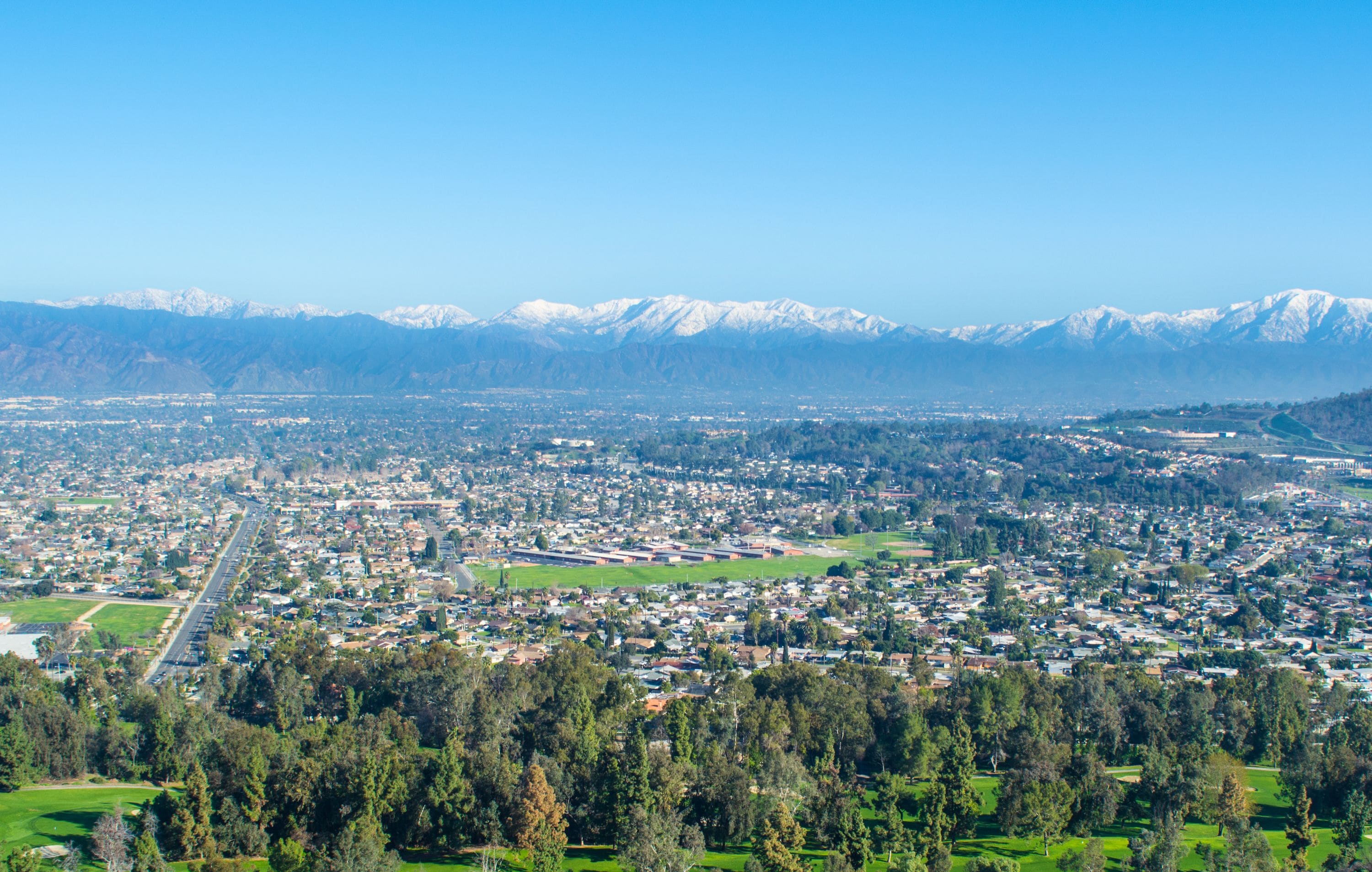 Una vista panoramica di una città con una strada centrale, zone residenziali e montagne innevate sullo sfondo.