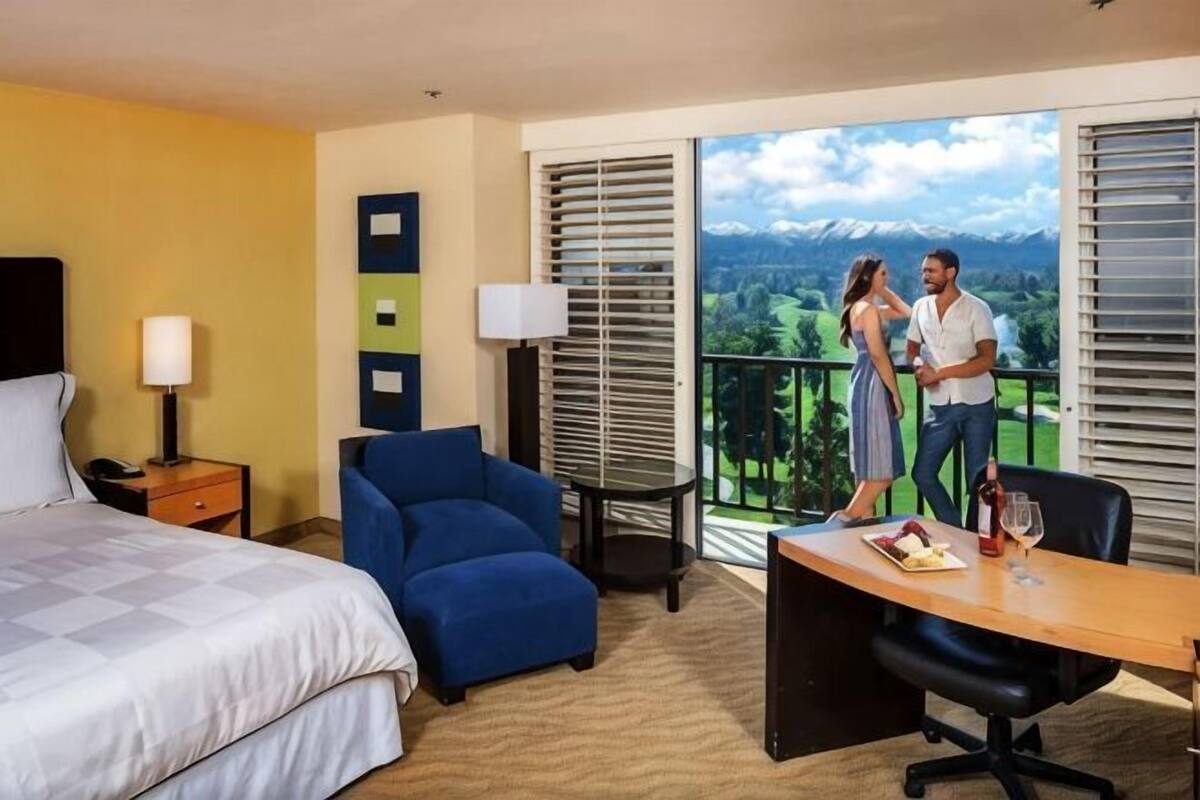 Une chambre d’hôtel avec un lit, un fauteuil bleu, un bureau avec une bouteille et un verre, et une vue sur les montagnes depuis le balcon.
