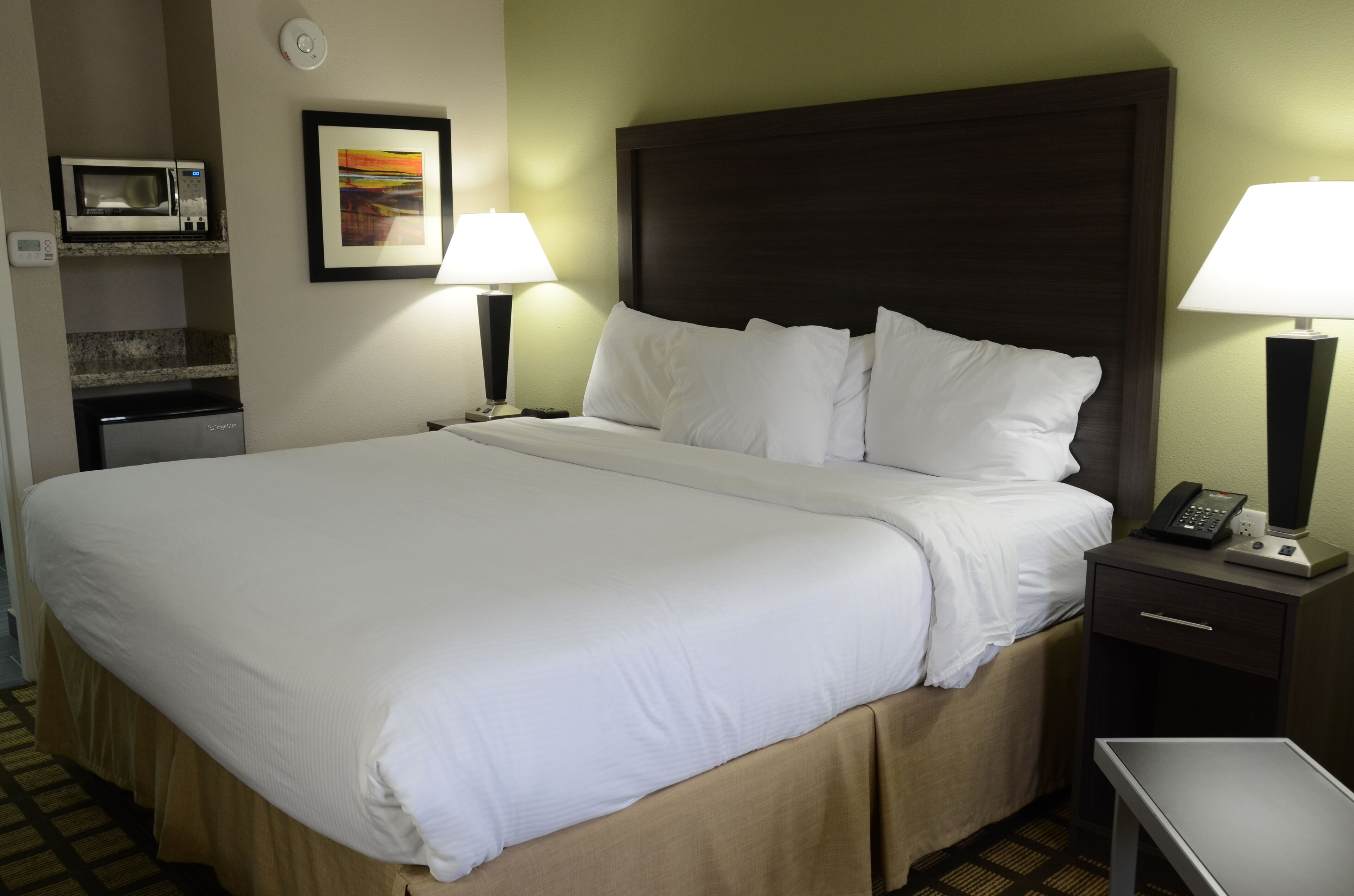 A hotel room with a large bed, two bedside tables with lamps, a microwave, a mini-fridge, and a framed picture on the wall.
