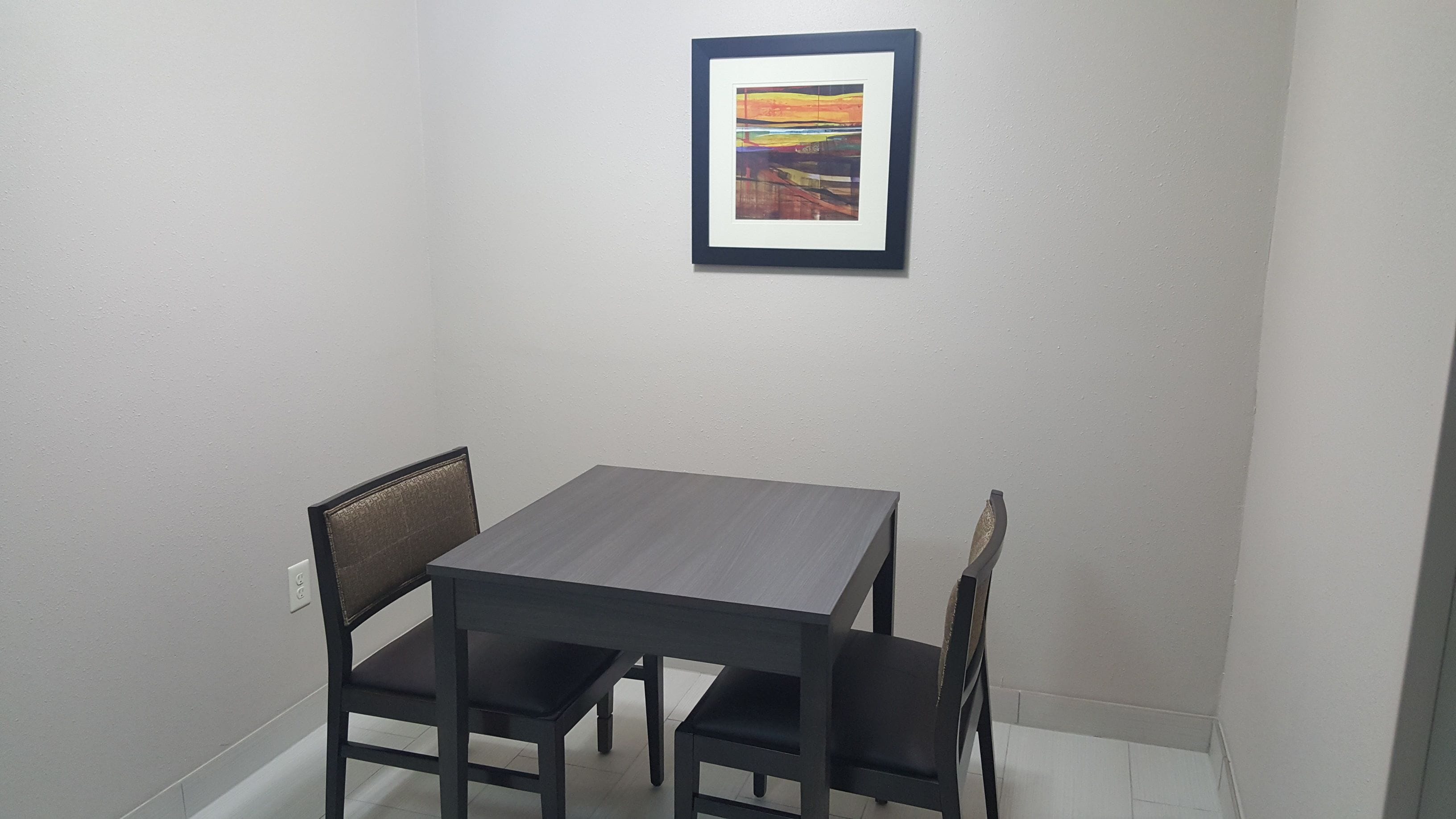 A small dining area with a table and four chairs, a framed picture on the wall, and a white tiled floor.