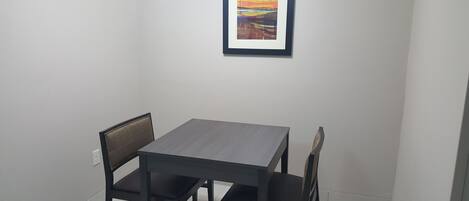 A small dining area with a table and four chairs, a framed picture on the wall, and a white tiled floor.